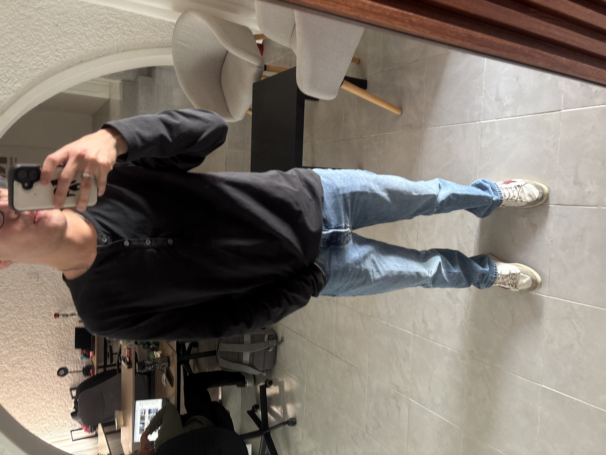 A person is captured in a mirror selfie, seen from the mid-torso down. They are wearing a black long-sleeved Henley shirt, light blue jeans, and white sneakers with red accents. Their face is largely obscured by the smartphone they are holding up to take the picture, though a glimpse of their profile and a ring on their hand are visible.
The scene is an indoor setting, likely in Puebla, Mexico, characterized by light-colored tiled flooring. In the immediate vicinity of the mirror, two modern, light grey armchairs with wooden legs are arranged around a dark, rectangular table. The mirror reflects a room beyond, which appears to be an office or workspace. This background shows several desks equipped with computer monitors (one displaying content), office chairs (one grey, one dark), and various items on the desks. A grey backpack rests on the floor near one of the chairs, and another person is partially visible seated at a desk. An arched opening or doorway leads to a section with a textured, possibly stucco, ceiling or wall. A wooden slatted structure frames the top edge of the mirror's reflection. The lighting suggests it is daytime. No discernible text is visible on any objects.