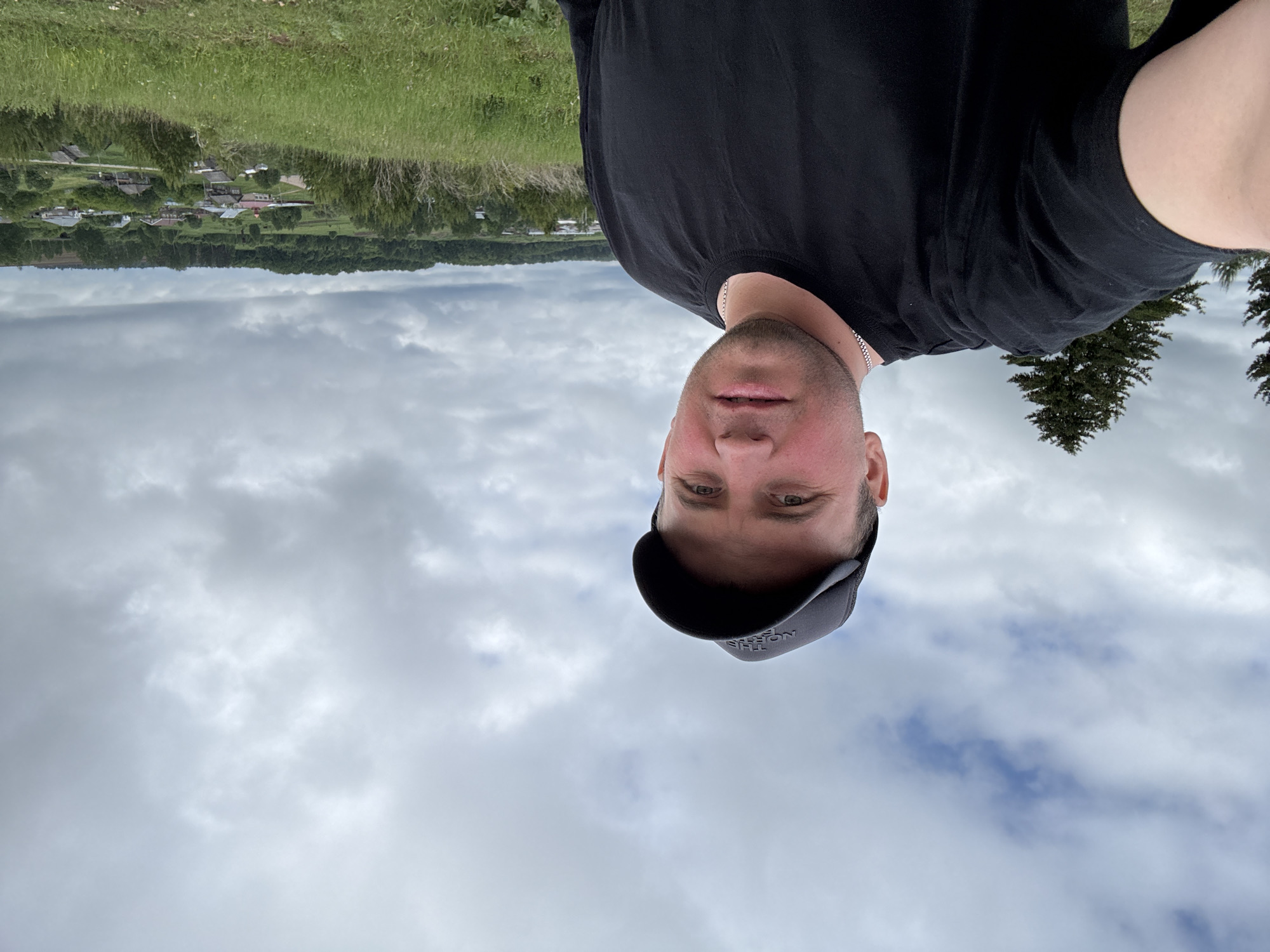 The image, rotated 90 degrees clockwise, is a selfie featuring a man in an outdoor, rural setting. The man, positioned in the foreground, looks directly at the camera with a neutral expression. He is wearing a black t-shirt, a silver chain necklace, and a dark baseball cap, which has a partially visible "THE NORTH FACE" logo on the brim.
Behind him, an expansive rural landscape unfolds under an overcast sky. A lush green grassy area occupies the immediate background, leading to a distant village nestled among trees. Several houses with varying roof colors, including red, are visible within the village. Beyond this, a dense line of dark green forest marks the horizon, possibly a distant ridge. The sky is filled with scattered, light grey clouds, indicating an overcast daytime. The scene suggests a tranquil, natural environment, consistent with the location context of Пычас, Russia.