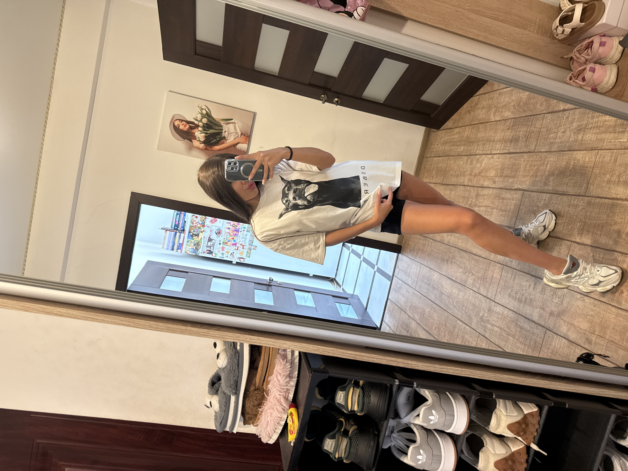 A young woman with long brown hair is standing in front of a full-length mirror, taking a selfie with her phone. She is wearing a white oversized t-shirt with a graphic of a dog on it, and black shorts. Her legs and feet are visible, and she is wearing white sneakers. The mirror reflects a room with a wooden floor and a dark brown door. To the left of the mirror, there is a black shoe rack with several pairs of shoes on it, including slippers, Adidas sneakers, and other athletic shoes. In the background, there is a poster of a woman holding flowers. The image is taken indoors, likely in a residential setting in Chișinău, Moldova.