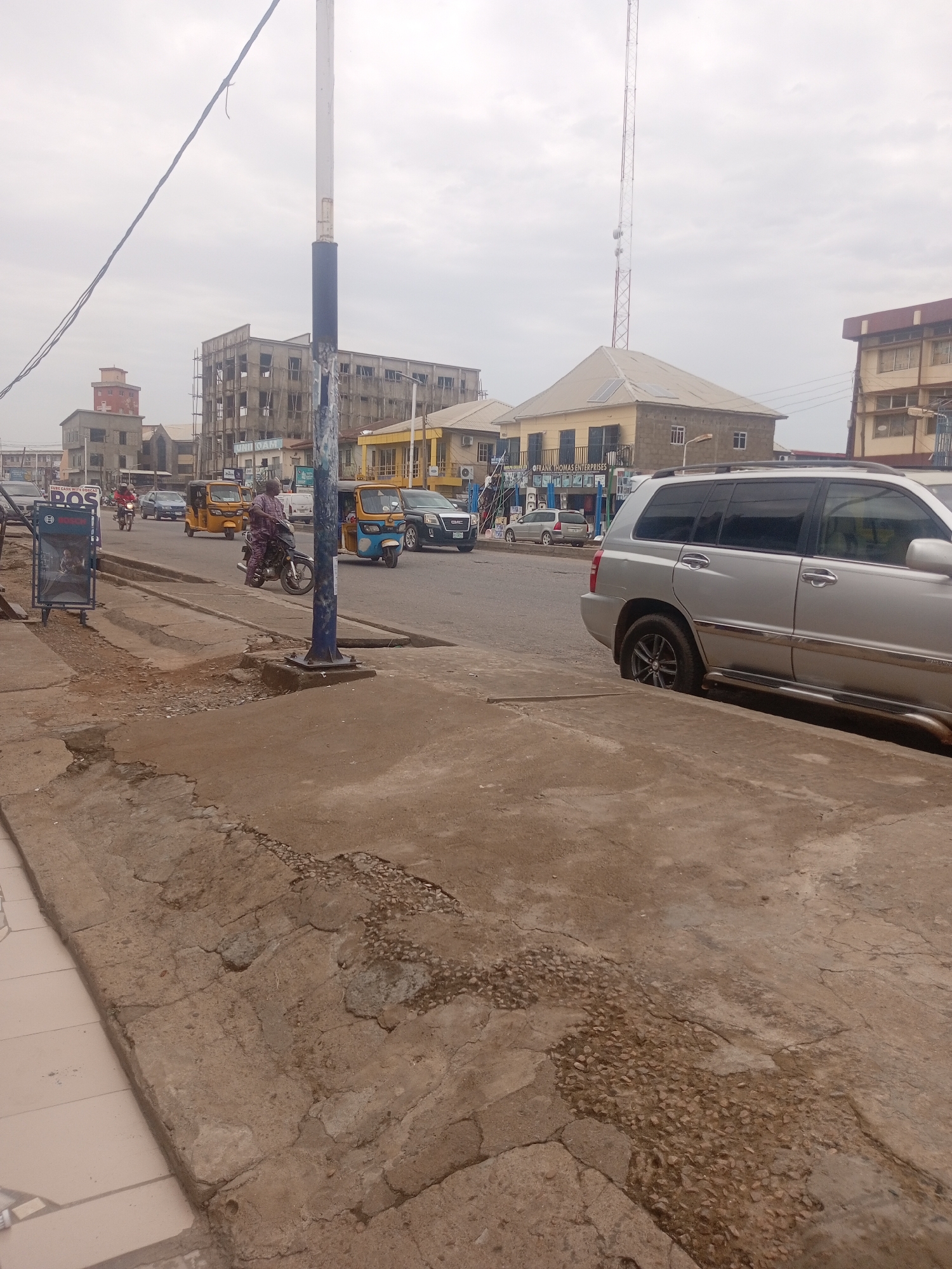 The image depicts a street scene in Sardauna, Nigeria, under an overcast sky. In the foreground, a cracked concrete sidewalk runs along the edge of a road. To the right, a silver SUV is parked or driving slowly. Across the street, a yellow auto-rickshaw, also known as a Keke Napep, is visible, as is a blue and yellow auto-rickshaw. A person is riding a motorcycle in the middle of the road, and another person is riding a motorcycle in the background. Several buildings line the street, including a multi-story structure under construction and other commercial buildings with various signage. A tall communication tower is prominent in the background. A blue and metal structure with signage, possibly a kiosk or advertisement display, is situated on the left side of the sidewalk. The overall environment suggests a moderately busy urban street.