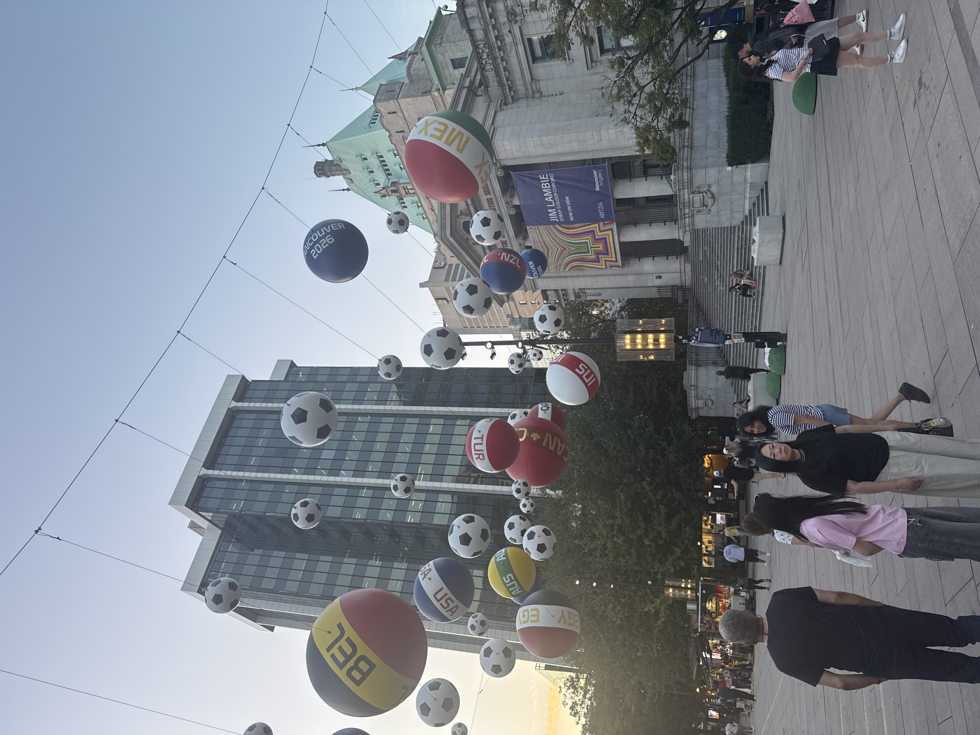 This is a medium-shot, eye-level photograph of a public plaza in Vancouver, Canada, likely in the downtown core, given the presence of the Vancouver Art Gallery and other large buildings. The image is taken during the late afternoon or early evening, as indicated by the soft, warm light and the clear sky.
The plaza is adorned with numerous large, inflatable spheres designed to resemble soccer balls, many of which are decorated with national flags or country abbreviations such as "BEL" (Belgium), "USA," "MEX" (Mexico), "TUR" (Turkey), "EGY" (Egypt), "AUS" (Australia), "NZL" (New Zealand), and "SUI" (Switzerland). Some of these are hung from wires overhead, creating a festive atmosphere. One large blue sphere prominently displays "VANCOUVER 2026."
In the background, the Vancouver Art Gallery, a grand neoclassical building with a green roof, is visible. To its left, a modern glass skyscraper stands tall. A banner hangs from the Art Gallery, advertising "JIM LAMBIE NOW ON VIEW."
In the foreground, several people are walking or standing in the plaza. On the left, a man wearing a black t-shirt and dark pants faces away from the camera. To his right, a woman in a pink t-shirt and jeans looks to her left. Next to her, a woman in a black t-shirt and light-colored pants is looking forward with a neutral expression. Further to the right, a woman in a striped t-shirt and black skirt is looking to her left, and a man in a grey t-shirt and shorts is walking away from the camera. Other individuals and groups are scattered throughout the plaza and on the steps leading up to the Art Gallery.
The ground of the plaza is paved with light-colored stone tiles, and trees line the edges of the scene. The overall impression is a lively urban space decorated for a significant event, possibly related to soccer or an international gathering.