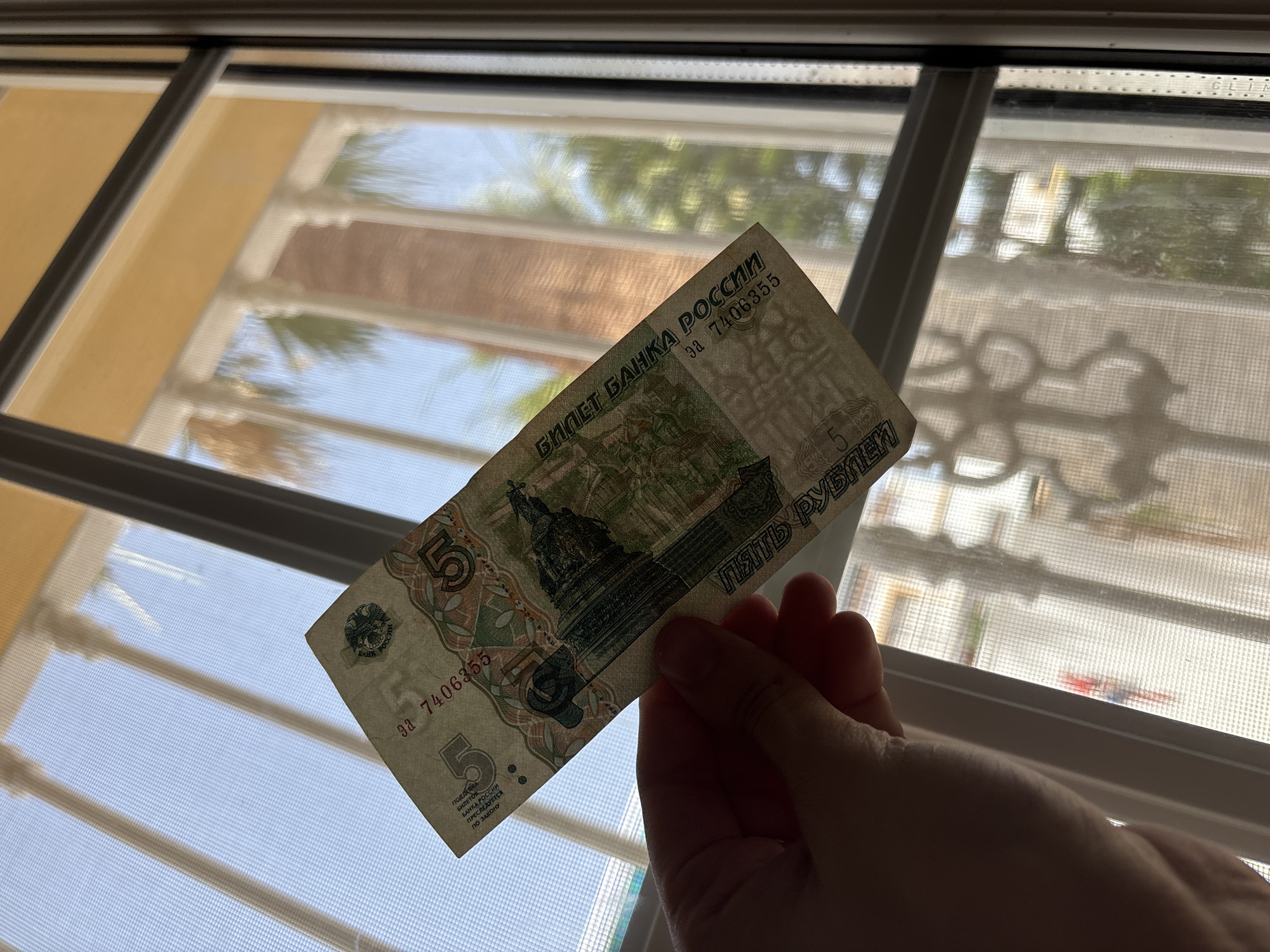 In the image, a person's hand holds a five-ruble Russian banknote up to a window. The banknote is partially translucent, allowing the bright sky and some greenery to be seen through it. The window appears to have a screen, and the blinds are partially open, casting striped shadows on the screen. Beyond the window, palm trees and a building with a yellow facade are visible, suggesting a sunny outdoor environment. The time of day is likely daytime, given the bright natural light. The banknote itself features the number "5" prominently, along with text in Russian that translates to "Bank of Russia Ticket" and "Five Rubles." There is also a warning about counterfeiting being punishable by law. The overall impression is a candid shot of someone examining a piece of currency against a backdrop of a warm, possibly Mediterranean, location.