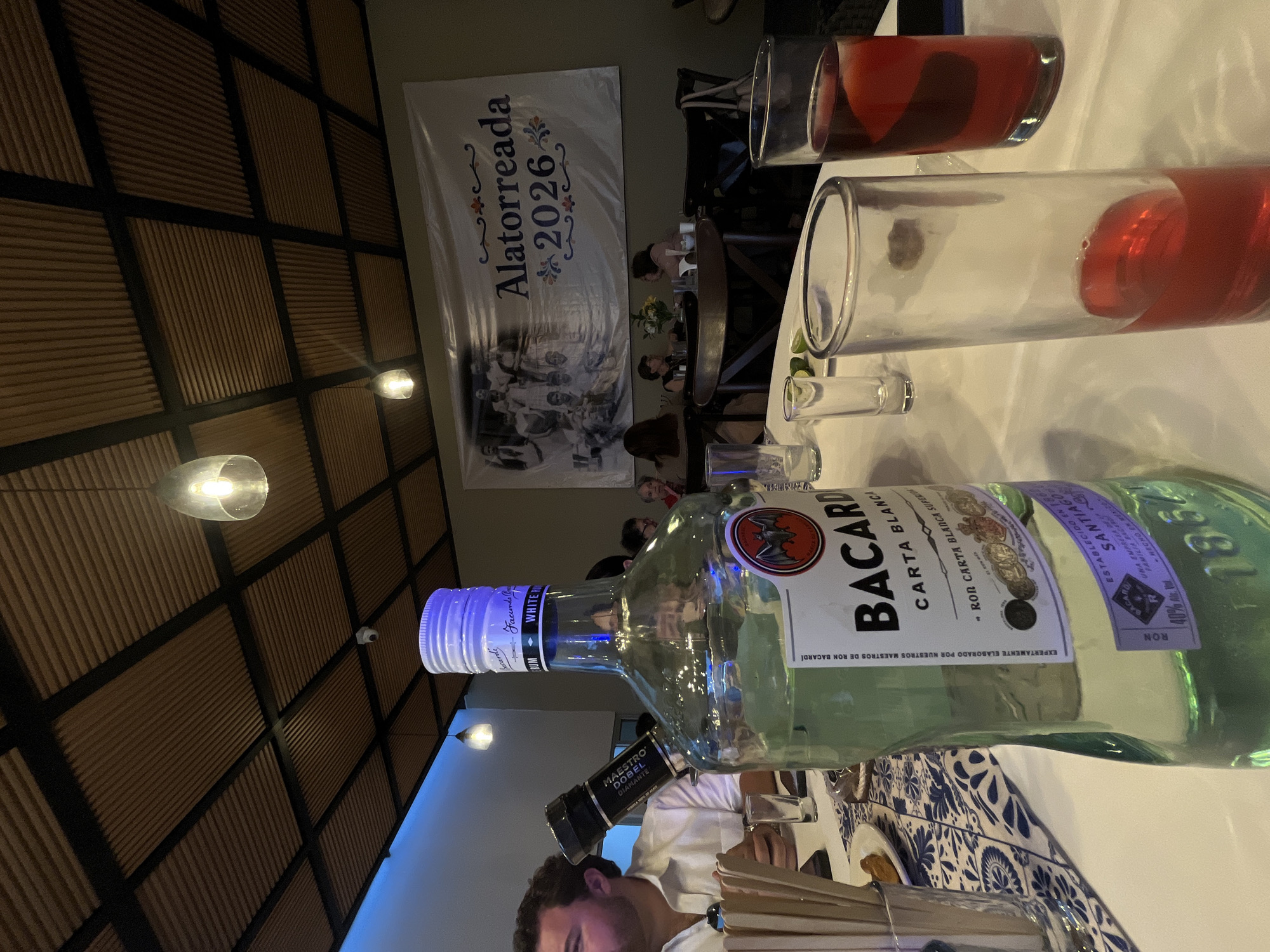Here is a description of the image:
**Location:** The photograph appears to be taken indoors, possibly in a restaurant, at an event in San Bernardino Tlaxcalancingo, Mexico, indicated by a banner in the background reading "Alatorreada 2026."
**People:** Several people are present, partially visible around tables. One man is prominently visible in the foreground, his face partially shown. There seem to be other people seated around tables in the background.
**Subjects/Objects:** The main objects are alcoholic beverages. There's a bottle of Bacardi Carta Blanca rum, and a bottle of Maestro Dobel Diamante. There are also several glasses, some containing a red liquid, likely a cocktail or mixed drink. Chopsticks are visible near the lower part of the frame. There is also a security camera visible on the ceiling.
**Actions/Interactions:** The image seems to capture a social gathering. It is unclear from the photo if any specific interactions are taking place.
**Environment:** The setting includes a dining table with a blue and white patterned tablecloth. The ceiling has a textured, grid-like design. There are hanging light fixtures.
**Overall Impression:** The photo depicts a gathering or celebration, possibly a private event. The presence of alcohol and the banner suggests a festive atmosphere.