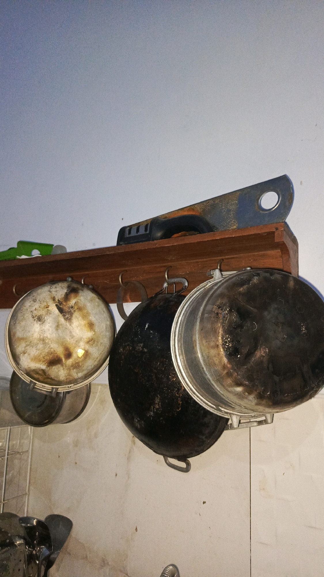 In a kitchen, a wooden shelf is mounted on a light-colored wall. The shelf holds three cooking pots of varying sizes. The pots are hanging from hooks on the underside of the shelf and appear to be well-used, with some discoloration and residue. On the shelf itself, a tool with a black handle and a metal blade is placed horizontally. Below the shelf, the tiled wall of the kitchen is visible, showing some grime and stains. The lighting in the scene is somewhat dim, casting faint shadows. There are no people or animals present in the image. The scene depicts a utilitarian kitchen space, suggesting a focus on practical use rather than aesthetics.