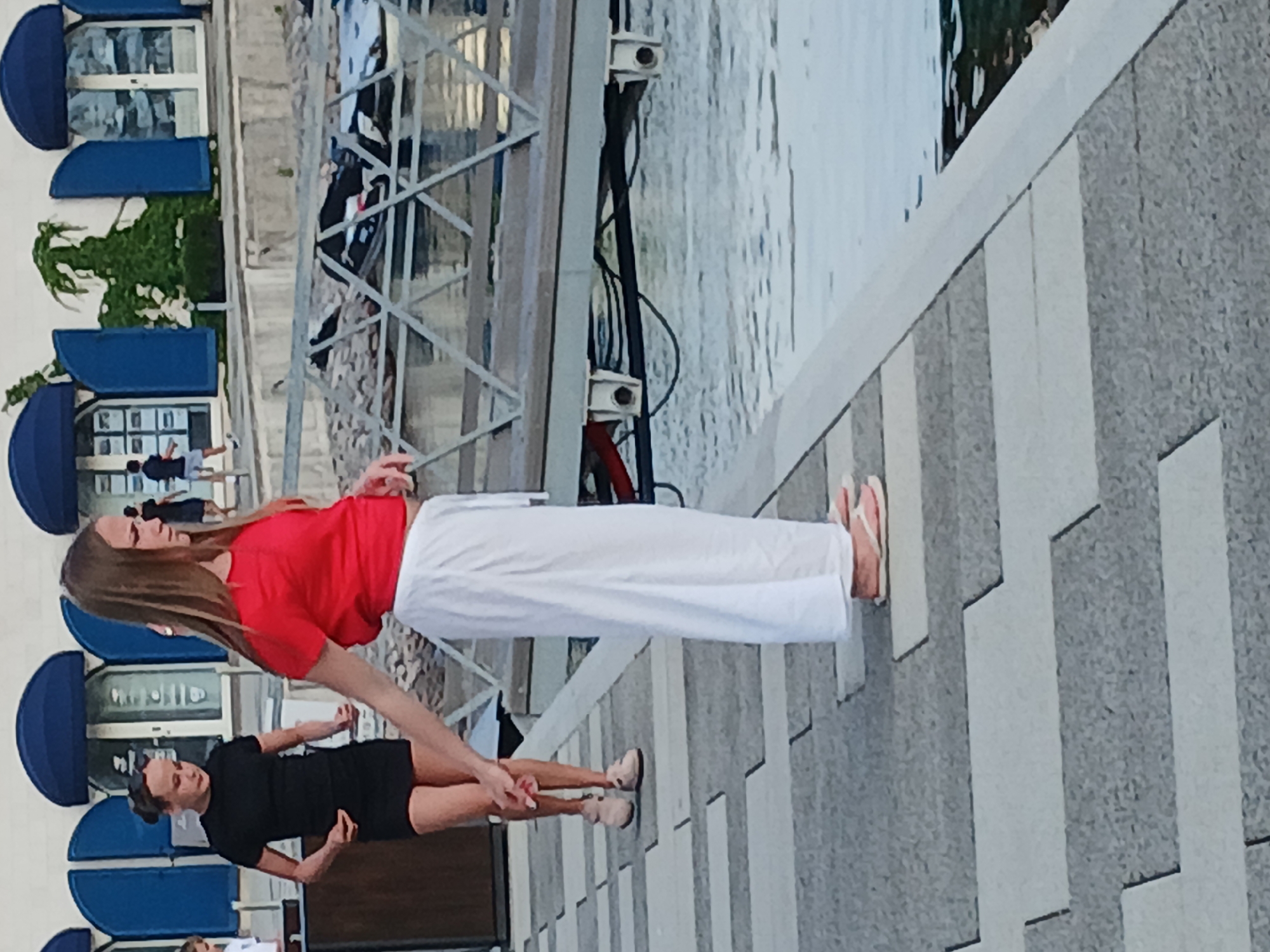 Two young females are on a paved walkway near a body of water. The female on the left is wearing a black t-shirt and black shorts, with her hair pulled back in a bun. The female on the right is wearing a red t-shirt and a long, flowing white skirt. She has long, light brown hair. They appear to be walking or standing together. In the background, there are buildings with blue awnings and a pier with boats. The walkway is made of large, rectangular grey and light grey pavers. The body of water appears to be calm with some reflections.
