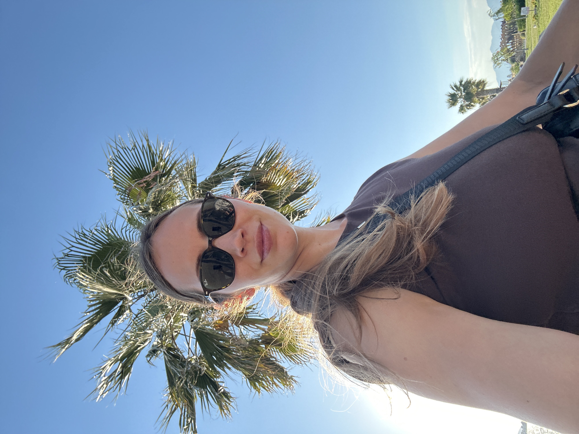 A woman with fair skin, brown hair, and sunglasses stands outdoors in Fethiye, Turkey. A palm tree is visible in the background. The sky is clear and blue. The woman is wearing a brown top and a black bag is slung over her shoulder. The angle of the photograph is low, looking up at the woman.