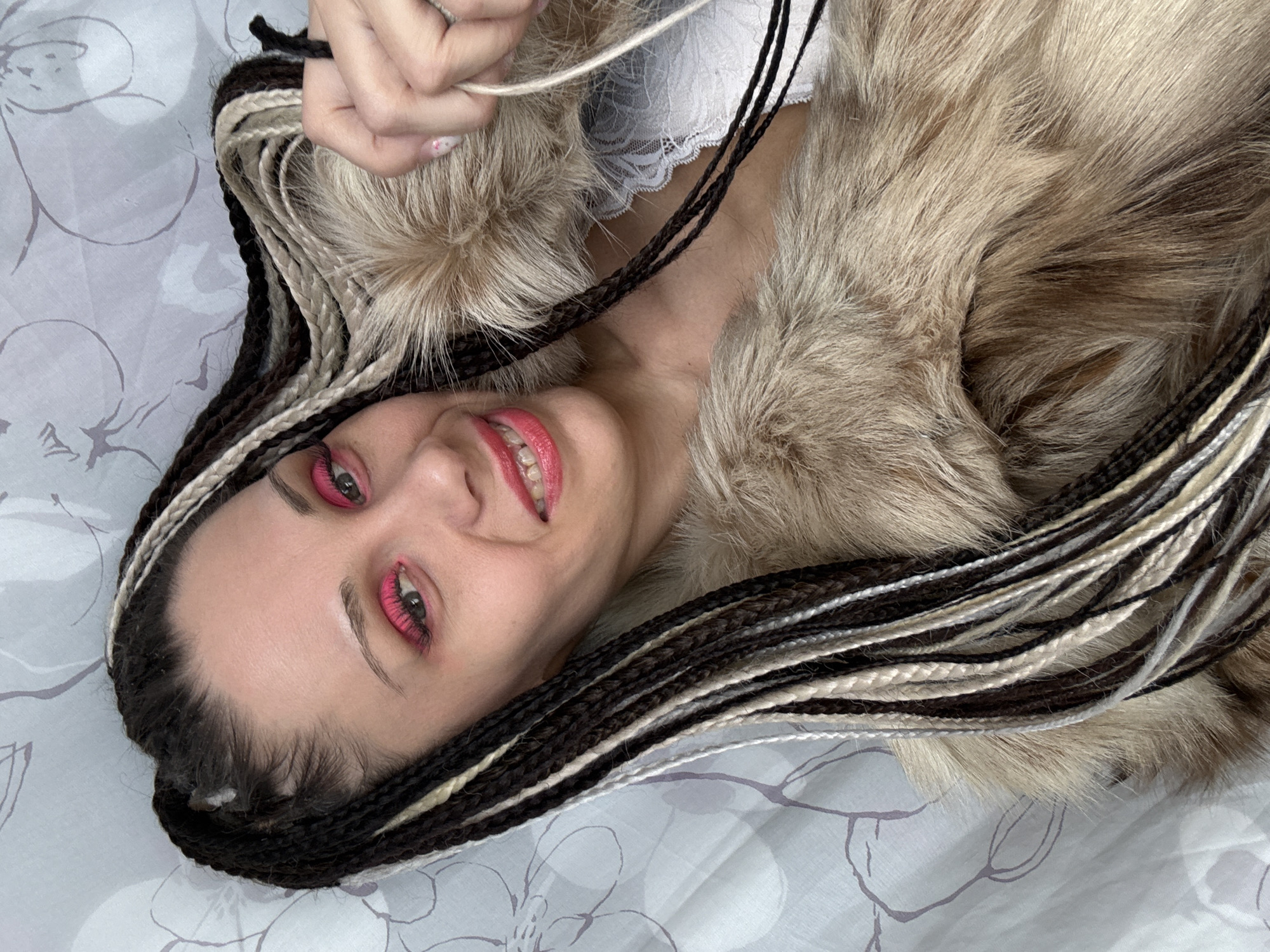 A woman with braided dark brown and light blonde hair lies on a white and gray patterned bedspread. She is wearing heavy bright pink eye makeup and lipstick. She is wearing a light-colored fur coat and a white lace top. Her hand is visible near the top of the frame, holding a strand of light-colored hair. No information indicates this photograph was taken in Rusanovo, Russia.
