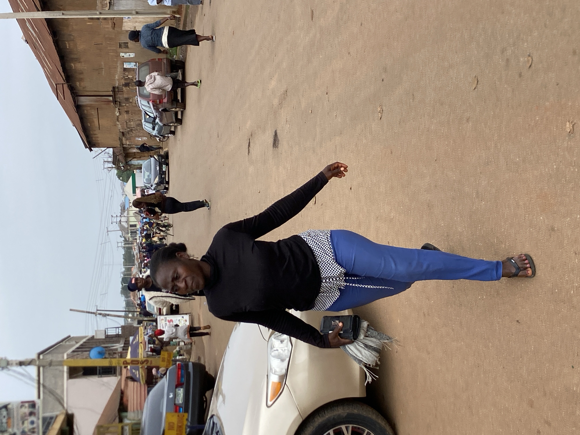 A woman in Bukuru, Nigeria, stands beside a beige car in a busy street scene. She wears a black long-sleeved shirt, blue pants with a white and black polka dot pattern at the waist, and black sandals. She holds a black wallet and a bundle of what appears to be grey cloths or paper. The street is lined with buildings, vehicles, and pedestrians. In the background, numerous motorcycles and people are visible, suggesting a crowded urban environment. The road surface appears to be reddish-brown.