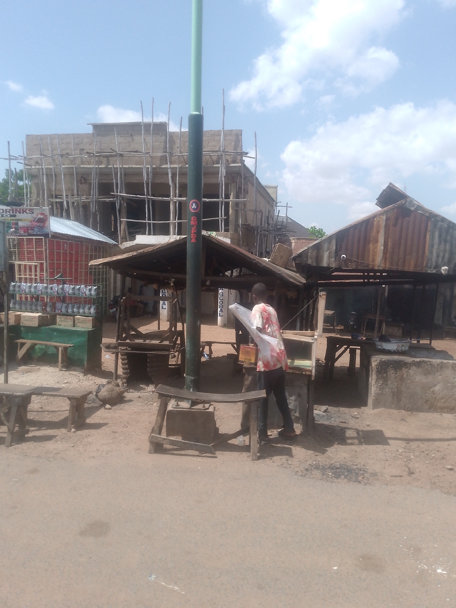 This image depicts an outdoor urban scene in Chabal, Nigeria, under a partly cloudy sky.
In the background, a multi-story building is under active construction, featuring concrete block walls and extensive scaffolding made of numerous wooden poles. To the left, a small, enclosed vendor stall with a corrugated metal roof displays "DRINKS" and "COCA-COLA" branding on a wall constructed from red crates. In front of this stall, various bottled beverages are arranged on shelves, and several stacks of uniform boxes, some labeled "AMMALCO," are visible.
Centrally positioned in the mid-ground is a light green utility pole with a darker base, featuring a circular red and white sticker that includes the letters "GMJPDA." Adjacent to this pole, a wooden-framed stall with a dark, possibly rusted metal or tarp roof, provides shade. Further to the right, a larger structure with a heavily rusted corrugated metal roof covers a darker interior where tables and indistinct figures are present.
In the mid-ground, a single individual is visible from the back, wearing a light-colored, patterned shirt (red, orange, and white hues) and dark trousers. This person is holding and manipulating a large, clear sheet of plastic material near a simple wooden stand.
The ground throughout the scene is dry, light brown earth or sand, with evidence of pathways. Several simple, unpainted wooden benches or stools are scattered in the foreground and mid-ground. A small, light-colored bowl rests on the ground near the right-most stall. The scene is illuminated by bright daylight.
