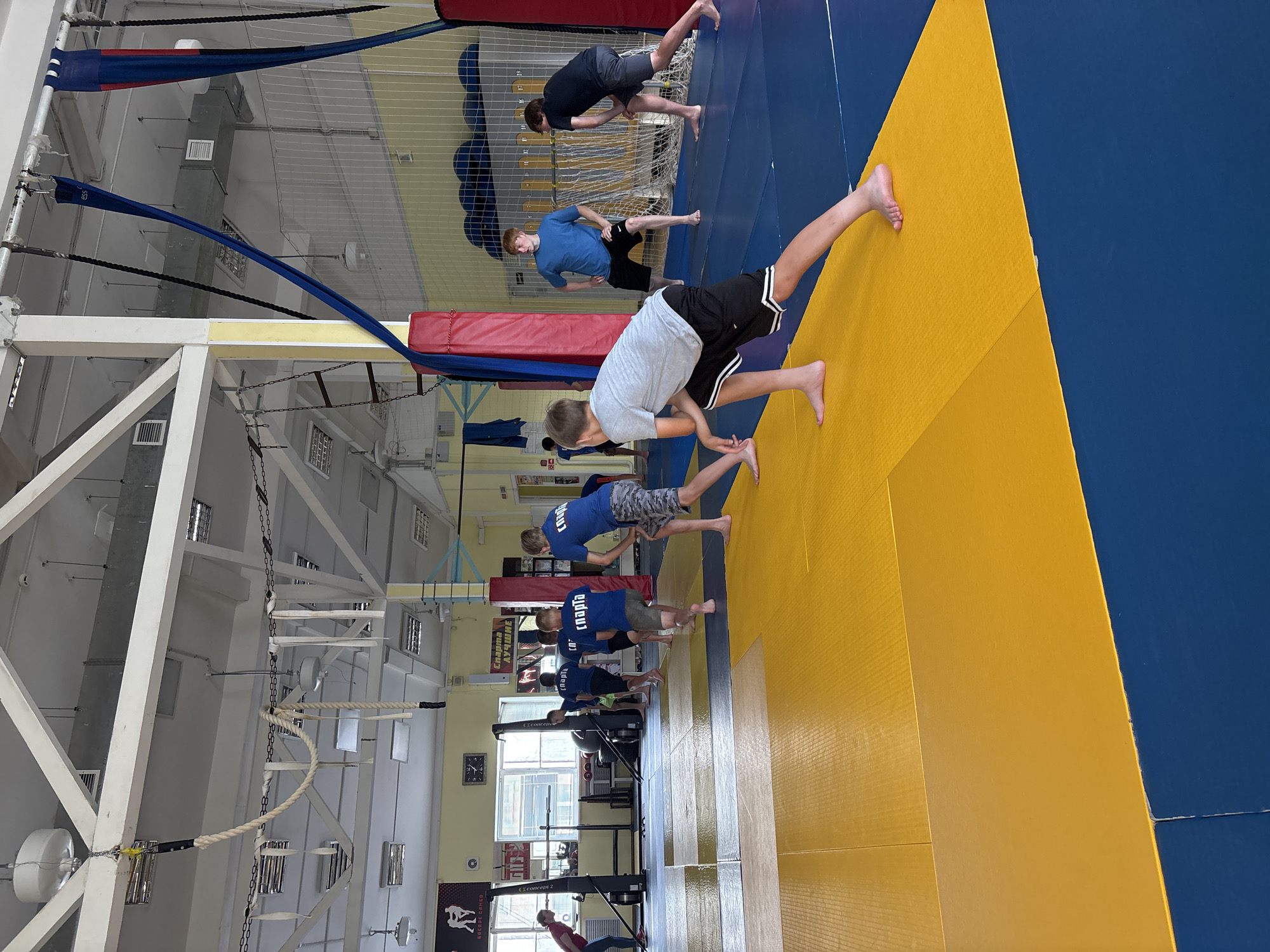 The image shows an indoor sports hall with a group of young people in athletic attire engaged in what appears to be a warm-up or training session. The floor is covered with blue and yellow mats, typical of a wrestling or martial arts facility. Several individuals are performing stretches, with their backs to the camera, wearing blue t-shirts with Russian lettering that likely indicates a sports club or team. There are also training apparatuses visible, including what seem to be rowing machines in the background. The ceiling has industrial lighting and a ventilation system, with ropes and fabric hangings suspended from the beams, suggesting an environment equipped for various sports activities. The overall scene is one of active physical training in a dedicated sports facility. The presence of Russian language on the t-shirts and the context of a sports hall suggests this location could be in Chaikovsky, Russia, a city known for its sporting infrastructure, particularly in winter sports, but also hosting facilities for other athletic disciplines.