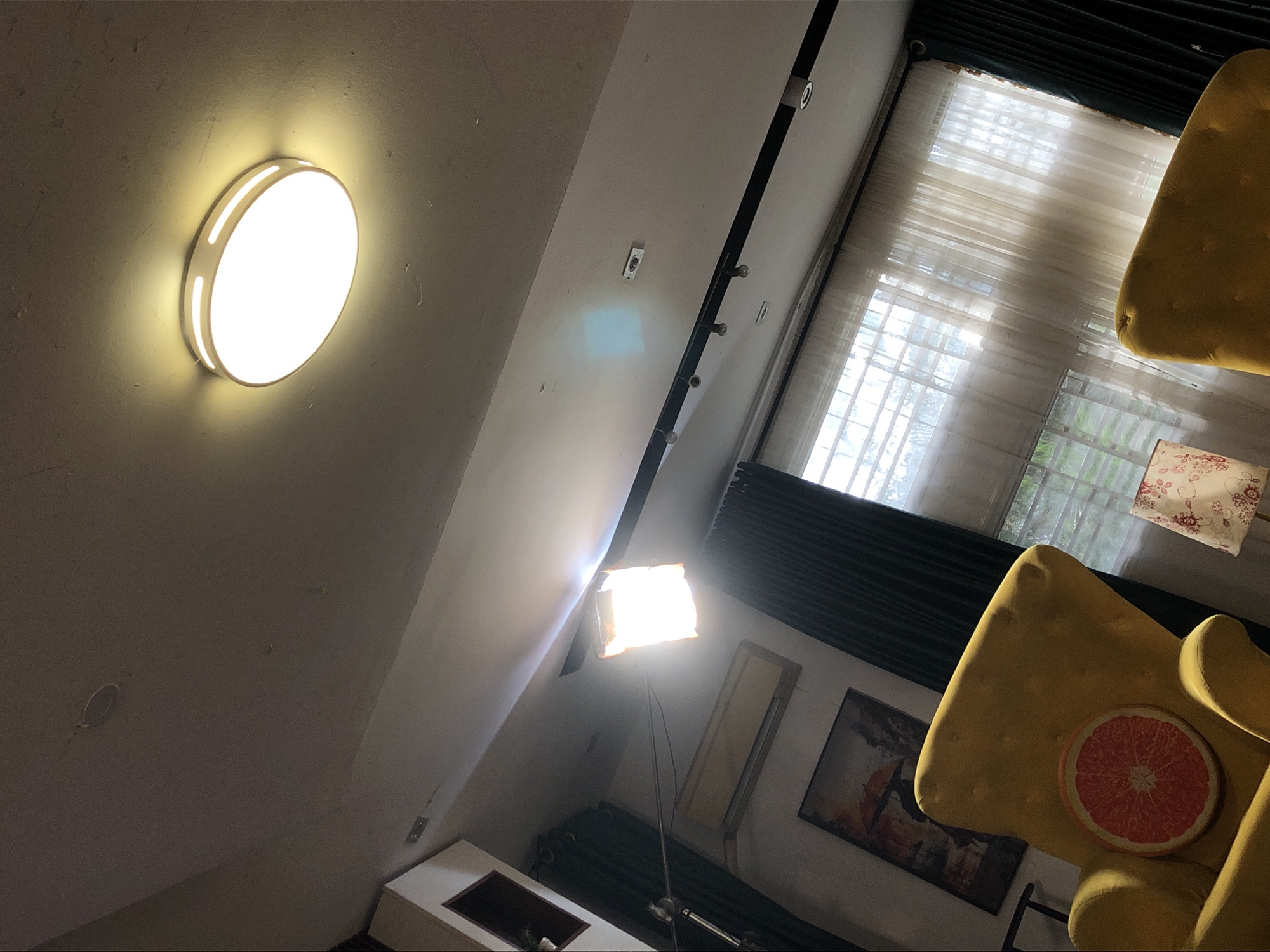 Here is a forensic description of the image:
The image is an interior shot taken from a low angle, looking upwards. The location appears to be inside a room.
A circular ceiling light fixture is visible on the left side of the image, attached to a light gray ceiling. A black curtain rail extends along the upper part of the scene. The wall has several electrical outlets.
To the right of the wall, there is a window covered with light curtains. Below the window, there's a dark gray radiator.
In the lower-right corner, a yellow armchair with a red grapefruit pillow can be seen.
A lamp on a stand is visible, casting a bright light towards the center of the image.
Below the lamp, there is a white shelf and some decor elements.
The painting is on the wall as well.
There are no people visible in the image.