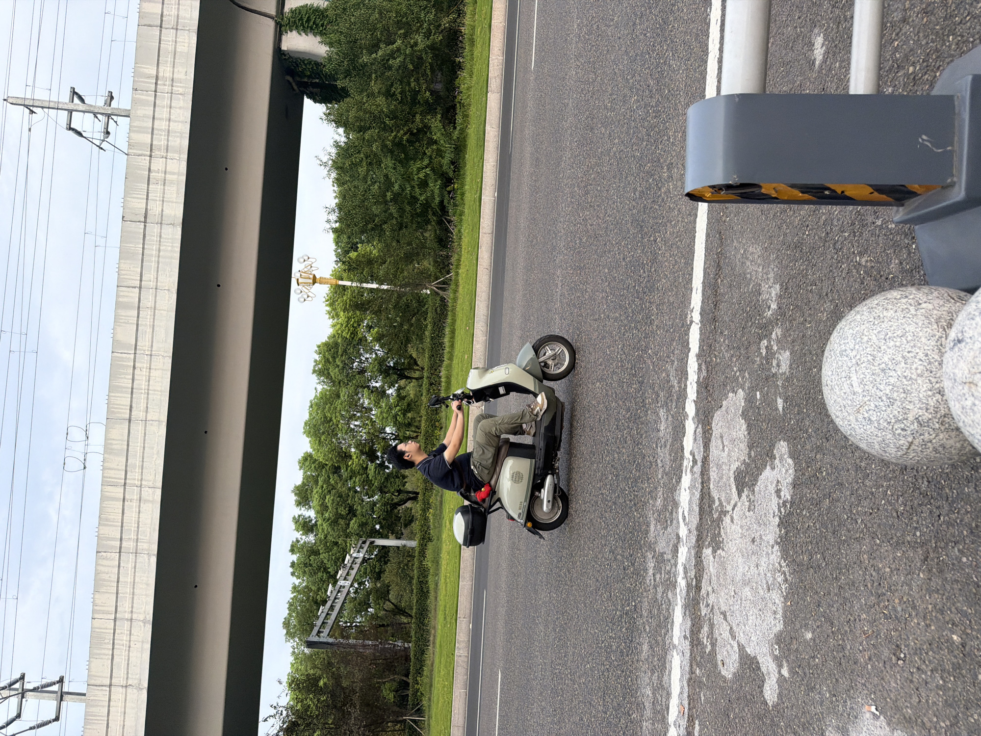 In Jiangjin, China, a man rides a light-colored electric scooter on a paved road. He is wearing glasses, a dark polo shirt, and khaki pants, with a black helmet box behind him on the scooter. The scooter has a white and light green body. The road has a white line dividing lanes. Overhead, a concrete overpass stretches across the scene, with power lines and poles visible. Lush green trees and vegetation line the side of the road, and a decorative lamppost stands to the left. In the foreground, a section of a gray barrier with yellow and black stripes and decorative spherical bollards are visible on the side of the road. The sky is overcast.