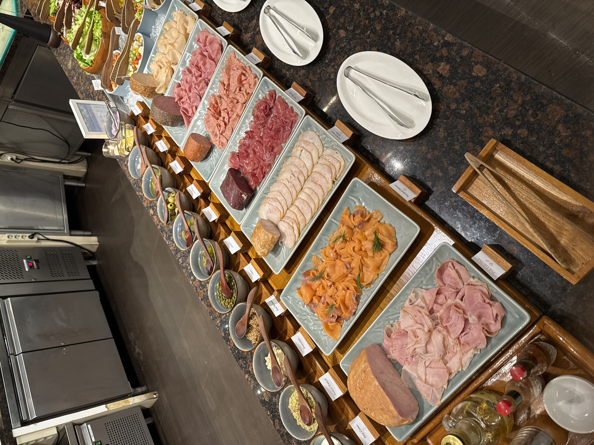 An overhead, eye-level shot captures a buffet spread at a hotel in Kuta Selatan, Indonesia. The counter is made of dark, speckled granite. In the foreground, a large wooden tray holds two small bottles of condiments, one clear with a gold cap and the other with a red cap, and a small white bowl. Next to it, a light blue rectangular tray holds thinly sliced pink ham, a larger piece of light pink ham, and a slice of dark pink meat. To the right of that, a matching tray displays thinly sliced pink deli meat, and a larger rectangular tray contains more sliced deli meats. Further to the right, a white oval plate holds a pair of silver tongs, and another similar plate below it also holds tongs.
The middle ground of the image features a light blue rectangular tray filled with smoked salmon, garnished with dill. To its left, several small grey bowls are filled with various condiments. One bowl contains a yellow condiment and is accompanied by a wooden spoon. Another contains a grainy brown substance and a wooden spoon. A third bowl holds a beige paste with a wooden spoon, and a fourth holds a pale green, chunky mixture with a wooden spoon. There are small white labels in front of each bowl. Further back, a large round wooden salad bowl is filled with leafy green lettuce. Next to it are several other rectangular light blue trays filled with various prepared foods, including what appears to be salads and deli meats. A round, dark loaf of cured meat sits on the counter. In the background, stainless steel kitchen appliances are visible, including refrigerators. The overall lighting is bright and even.