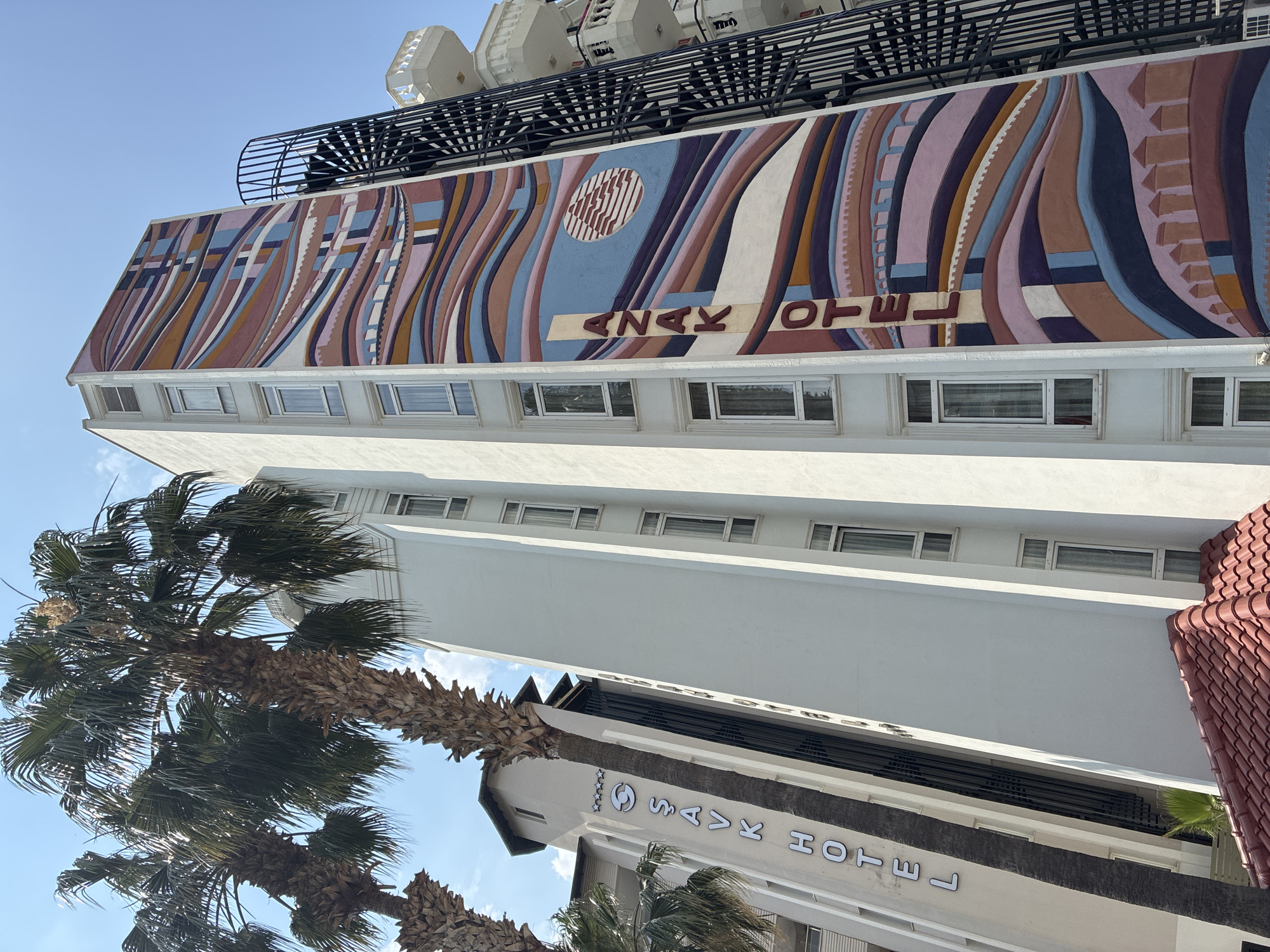 This is a slightly low-angle outdoor shot of two hotels in Alanya, Turkey, on a clear, sunny day. The hotel on the left is named "ŞAVK HOTEL" and has a stylized "S" logo above the name. It is a white building with a peaked roof and palm trees in front. The hotel on the right is taller and features a vibrant, wavy mural that spans its side, with the name "ANAK OTEL" vertically oriented on the mural. The mural uses a palette of pinks, blues, purples, oranges, and brown, with abstract geometric shapes and what appear to be stylized buildings at the bottom. A black spiral staircase is attached to the side of the "ANAK OTEL", and a white balcony structure is visible above it. The sky is a clear blue.