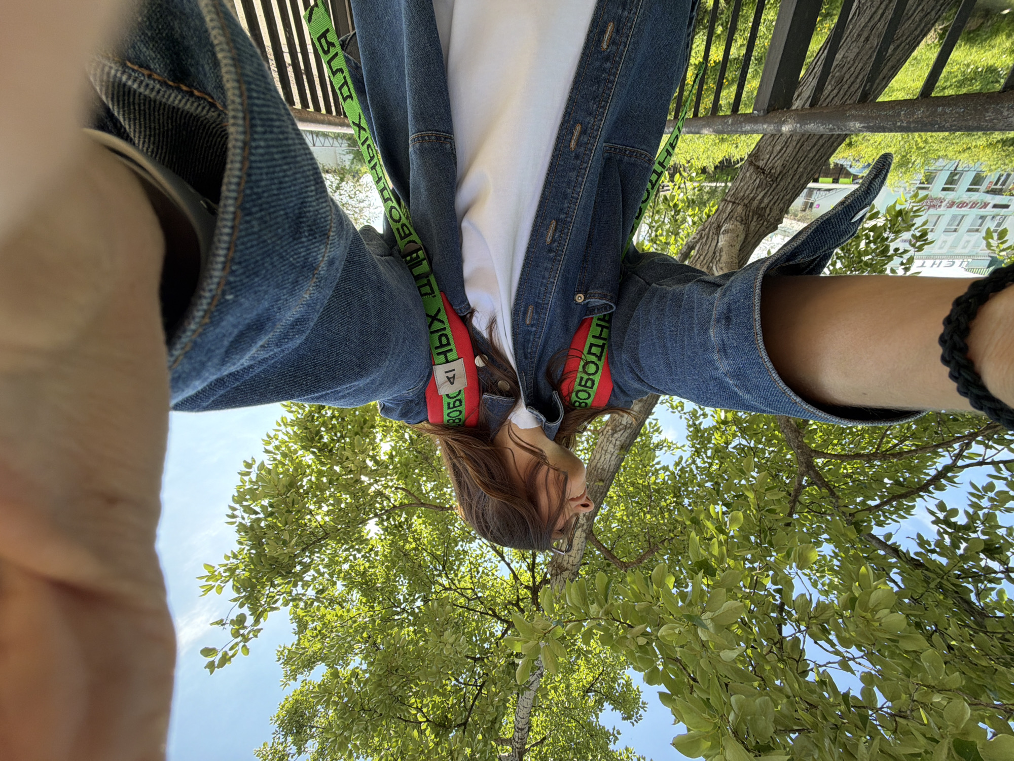 A low-angle, wide shot captures a person from the chest up, holding a camera out for a selfie. The person, with long brown hair, wears a denim jacket over a white t-shirt and a backpack with red straps adorned with green text. The background features a tree with bright green leaves against a clear blue sky, and a railing runs horizontally across the lower part of the frame. In the distance, on the left side of the image, a light blue building with white trim and several windows is visible. The scene appears to be outdoors, in an urban setting. It is daytime, and the sun is shining. The image is shot from a ground-level perspective, looking up. The building in the background is identified as being in the city of Birobidzhan, Russia.