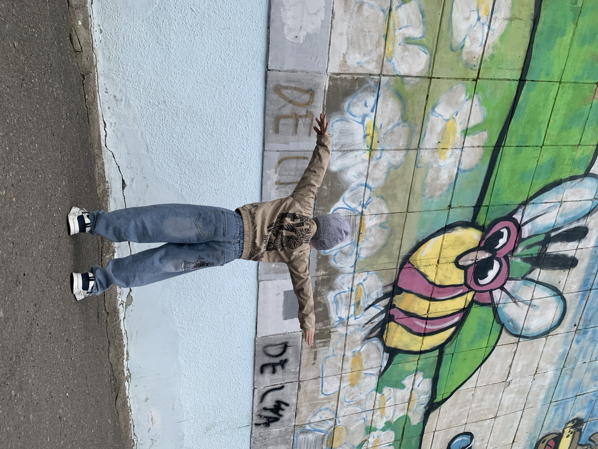 A full-body shot of a young person stands with their arms outstretched, creating a T-shape, in front of a colorful mural, positioned in the Russian city of Severodvinsk. The young person is dressed in blue jeans with graphics on the back of one leg, a light brown jacket with a printed graphic on the back, a dark beanie, and dark and white athletic shoes. They are standing at the intersection of a sidewalk and a painted wall. The wall appears to be decorated with a mural featuring a large bee with pink stripes. The wall is made up of square tiles. Graffiti writing is present on the wall, near the left side. The ground on the left side is asphalt and shows signs of wear. The wall is painted light blue.