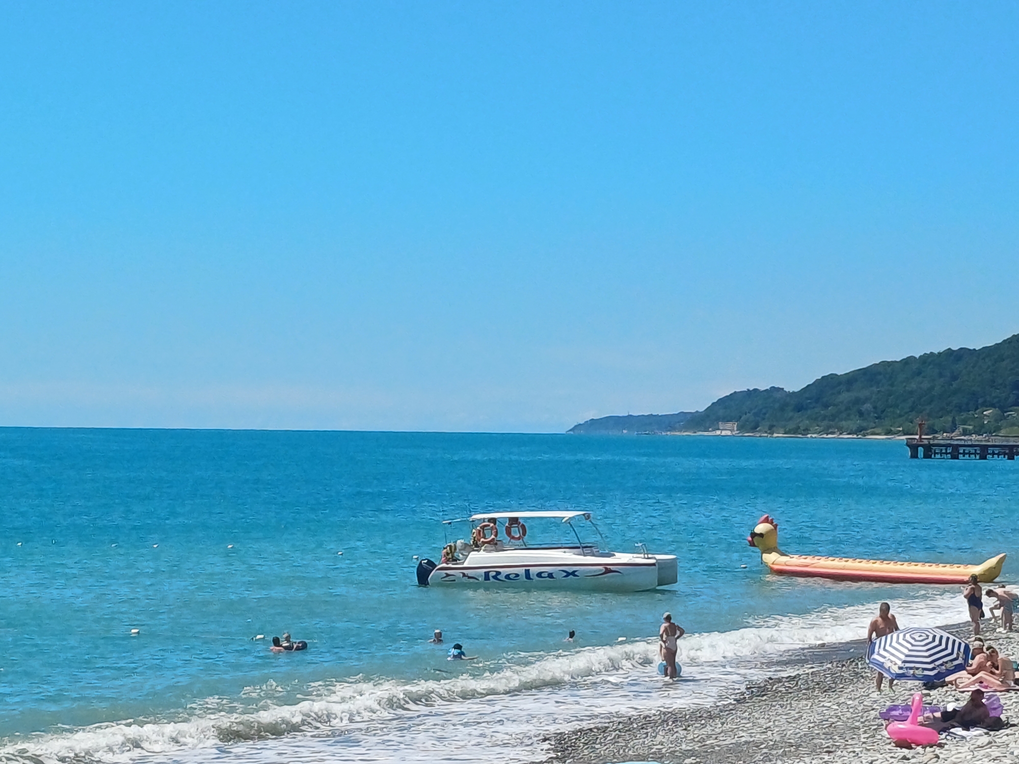 This is a photograph of a beach scene on a clear, sunny day in Sochi, Russia. The turquoise waters of the Black Sea are calm, with gentle waves lapping at the pebbled shore. In the water, a white speedboat with the word "Relax" emblazoned on its side is anchored. Several people are in the water, swimming and playing. To the right of the speedboat, a large, inflatable yellow banana-shaped toy floats near the shore.
On the beach, several people are sunbathing under umbrellas and towels. A striped umbrella is open, and individuals are seated or lying on the sand. Pink inflatable toys are scattered on the beach. In the background, a lush green hillside rises from the sea, with a long pier extending into the water. The sky is a clear, bright blue, indicating fair weather.