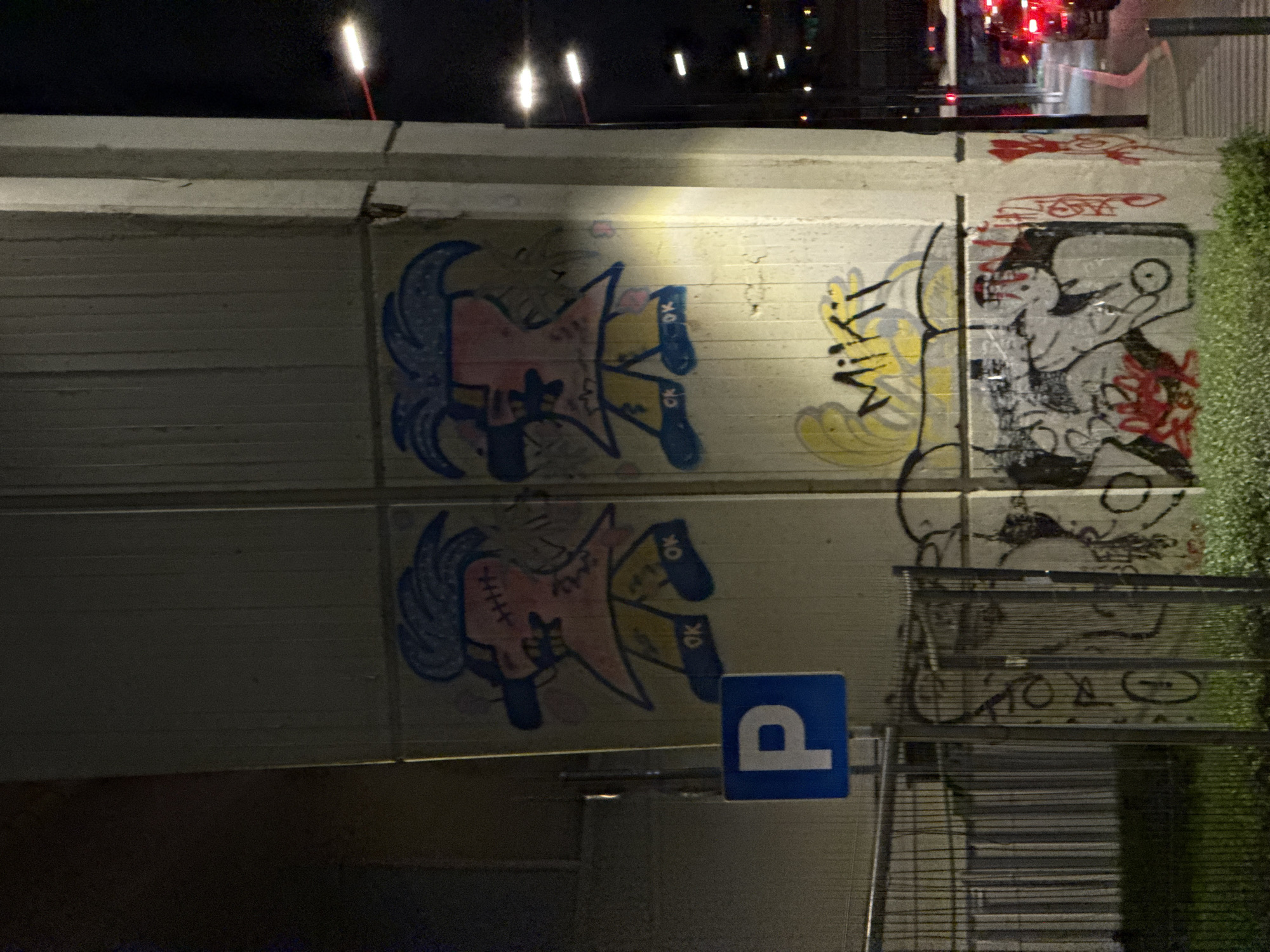This night-time photograph, taken in Belgrade, Serbia, captures a section of a concrete urban wall adorned with various graffiti and tagging. The main artwork on the left features two vertically stacked, identical stylized characters. Each character has a pink face with stitch marks, blue hair, and a yellow body or clothing, wearing blue boots with the text "OK" visible on them.
To the right of these characters, another graffiti piece includes distinctive yellow lettering that appears to spell "Miki," accompanied by a more abstract black and white creature-like figure with scattered red markings and outlines. Further right, the wall shows additional black outlines and red scribbled tags.
The scene is set at night, with a dark sky illuminated by several bright, elongated artificial lights in the upper portion of the image, and some distant red lights possibly from traffic or buildings. In the lower part of the image, a blue square sign with a white capital "P" (indicating parking) is attached to a pole. A dark metal fence or barrier runs along the bottom, partially covered by green foliage on the right side. The concrete wall itself appears to be part of an urban structure, such as an underpass or a roadside barrier.