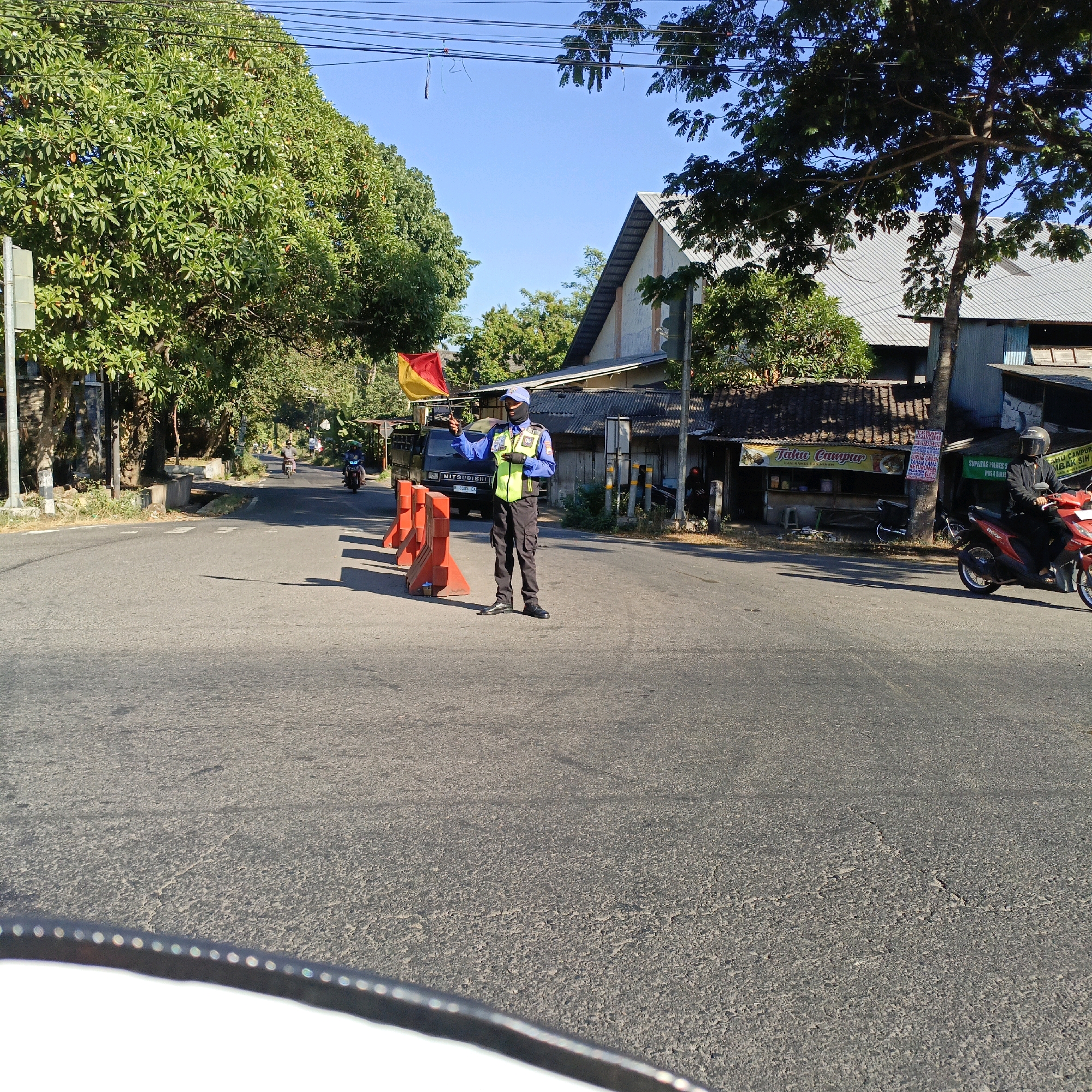 The image depicts a traffic director, wearing a blue cap, black face mask, blue long-sleeved shirt, and a reflective vest, holding up a red and yellow flag on a street in Salatiga, Indonesia. The traffic director stands on the right side of the road, with traffic cones placed in front of them. Behind the traffic director is a black Mitsubishi van. To the right of the road, there are buildings and trees. A red motorcycle with a rider wearing a black helmet is also visible on the right side of the road. The road appears to be a two-lane street with asphalt pavement. The sky is clear and blue, indicating a sunny day. The environment suggests a typical urban or semi-urban street scene in Indonesia.
