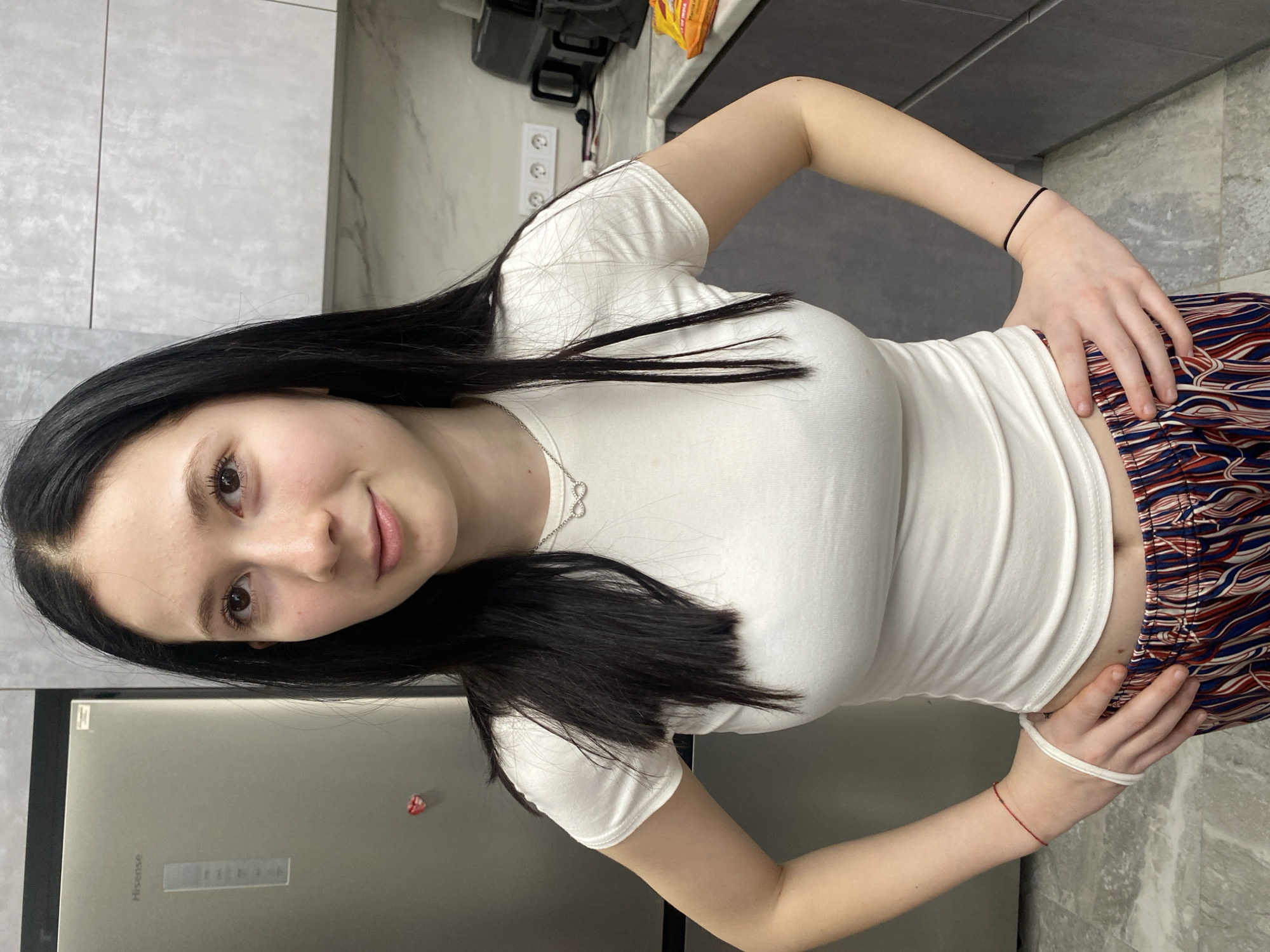 A young woman with long, straight dark hair, light complexion, and dark eyes is depicted standing indoors, likely in a kitchen, posing for the camera. She is looking directly at the viewer with a gentle, faint smile. She wears a fitted white short-sleeved t-shirt that slightly exposes her midriff, paired with loose-fitting, high-waisted pants featuring a bold, wavy geometric pattern in red, white, blue, and brown. Her hands are placed on her hips.
She accessorizes with a silver infinity symbol pendant necklace. On her right wrist, she wears a black hair tie or simple bracelet, and on her left wrist, she has a red string bracelet alongside a white hair tie or band.
The background indicates a modern kitchen setting. To her left, a stainless steel refrigerator with the "Hisense" brand logo is visible, adorned with a small red and white magnet. The wall behind her features large, light gray or white tiles, and a dark gray countertop is present. Further back on the counter, a dark appliance and an orange packet can be seen. Electrical outlets with three sockets are mounted on the tiled wall. The floor appears to be light-colored tiles. The indoor lighting suggests the photograph was taken during daytime or in a well-lit evening. The overall scene reflects a contemporary home environment in Izhevsk, Russia.