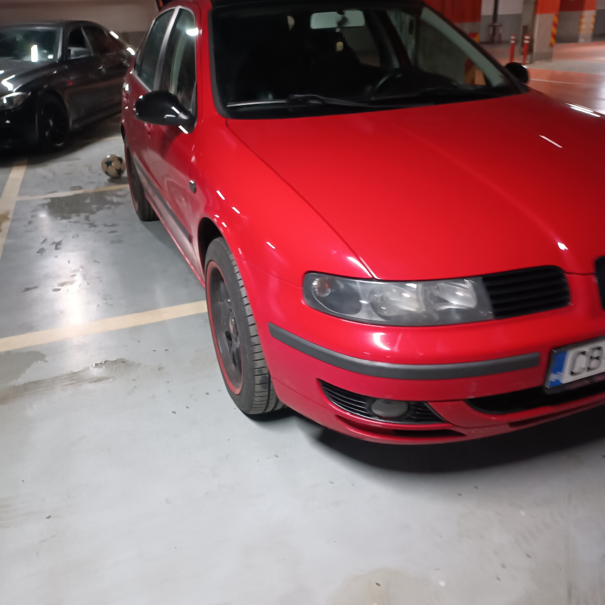 The image captures a scene in an indoor parking garage, likely in Sofia, Bulgaria. The primary subject is a vibrant red car, appearing to be a first-generation Seat Leon, parked prominently in the foreground. It features black alloy wheels accented with a red stripe, darkened headlights, and black side mirrors. A black protective strip runs across its front bumper. A partially visible European-style license plate on the front displays "CB," which is a common regional code for Sofia, Bulgaria.
Behind the red car and slightly to its left, a dark, likely black, car is partially visible, also parked within the garage. Between the two vehicles, resting on the concrete floor, is a soccer ball with a classic white and black panel design. The floor is light grey concrete with yellow parking lines, and some wet patches are visible, possibly from leaks or recent cleaning. The overall lighting suggests an artificial indoor environment, typical of a parking facility, with no indication of outdoor weather or time of day. No people are visible in the frame, and there are no discernible activities taking place beyond the static parking of the cars.