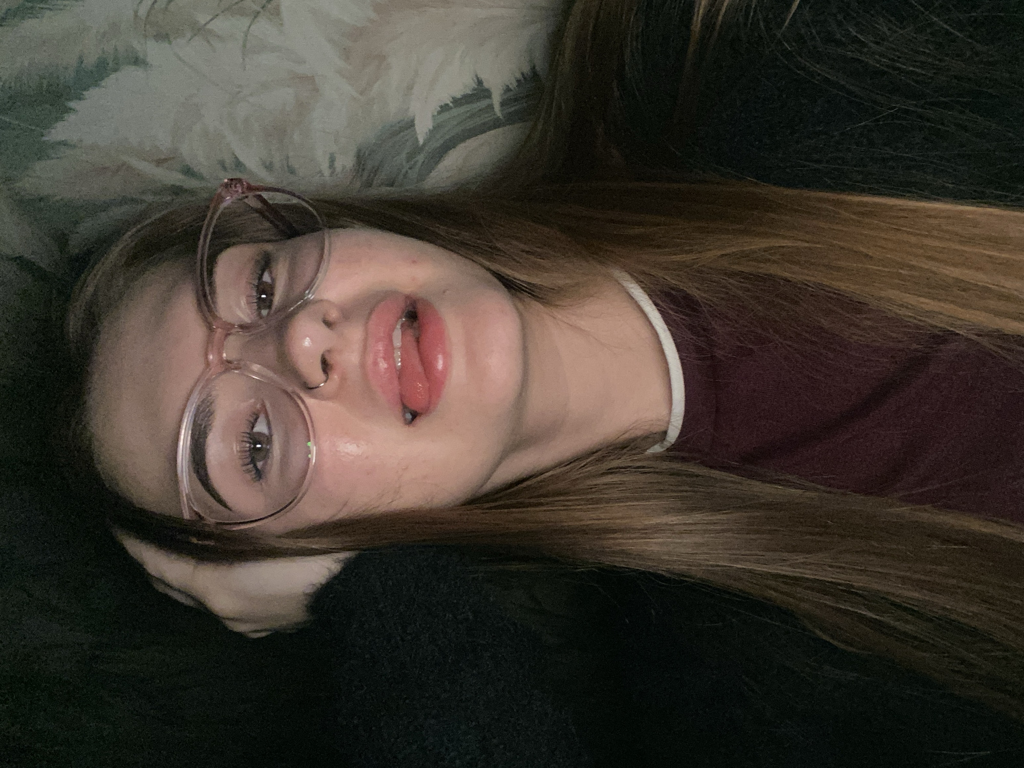 An indoor, eye-level shot captures a young person, likely female, lying down casually. Her head is turned to face the camera, with her long, light brown hair spread out around her. She wears light pink, clear-framed glasses, through which one eye is visible, looking directly at the viewer. Her mouth is slightly open, lips appearing glossy, with her tongue partially extended. A small silver nose ring adorns her left nostril. She is dressed in a dark red or maroon top with a white collar trim, and a dark, possibly black, textured fabric (perhaps a sweater or blanket) is visible beneath her chin and arm. The background features a patterned fabric in shades of light green, white, and cream, suggesting upholstery or a decorative textile. The lighting is soft, indicative of an indoor setting, and the person's expression appears relaxed and playful. There is no visible text.