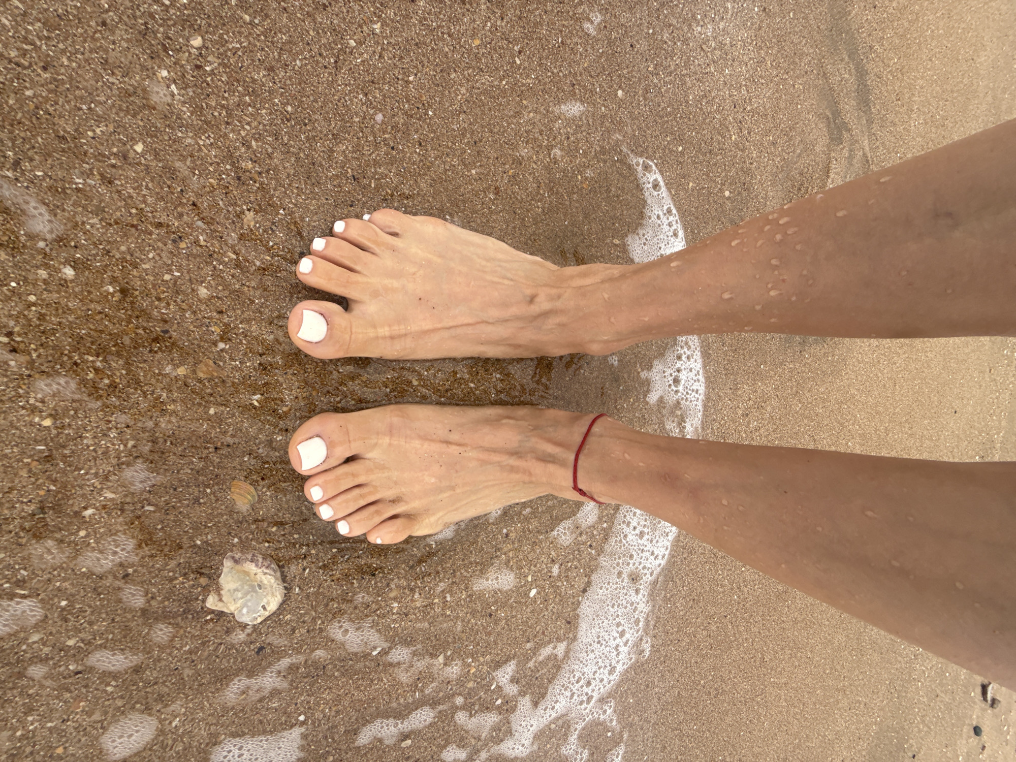 The image shows a pair of bare feet with white nail polish standing in wet sand on a beach. Water from the sea is washing over the feet, creating small waves and foam. The sand is a light brown color and appears to be composed of fine grains with some small shells and pebbles. The feet are positioned in the center of the frame, with the legs extending upwards out of view. A thin red string is visible around the left ankle. In the lower right corner of the image, a jellyfish is partially submerged in the sand. The lighting suggests it is daytime. The location is identified as the city of Kerch, Ukraine.