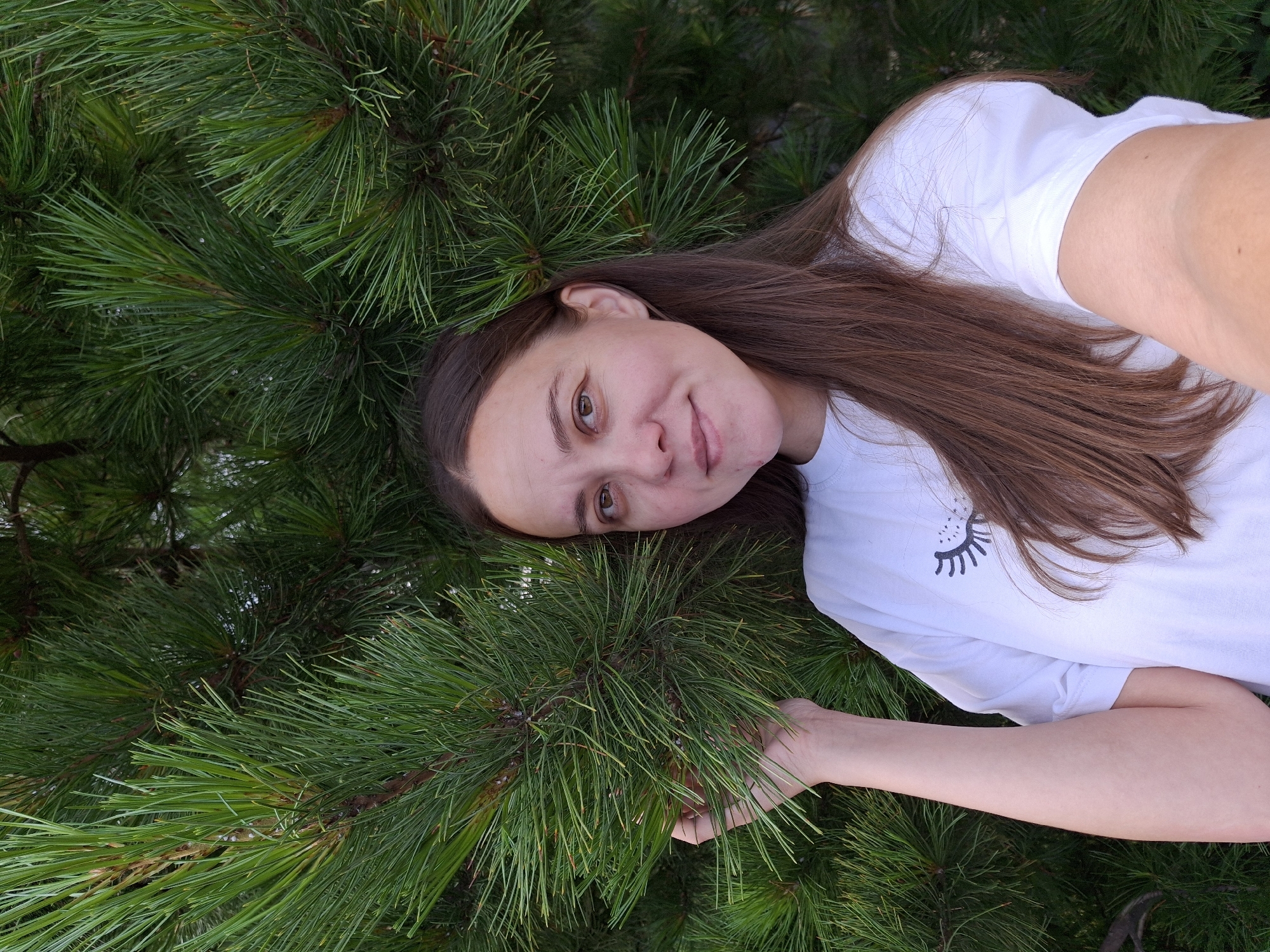 A close-up, rotated portrait shows a young woman positioned amidst dense, vibrant green pine branches and needles, suggesting an outdoor natural setting in Chaikovsky, Russia. The woman, with long, straight, dark brown hair and fair skin, gazes towards the camera with brown eyes and a subtle, calm smile. She wears a plain white short-sleeved t-shirt featuring a stylized black graphic resembling a sleepy eye or eyelashes on the left side of her chest. Her right arm is extended, with her hand gently resting on or holding a cluster of the pine needles. The lighting is bright and even, indicating daytime. The scene conveys a relaxed and serene mood, with the woman appearing connected to her natural surroundings.