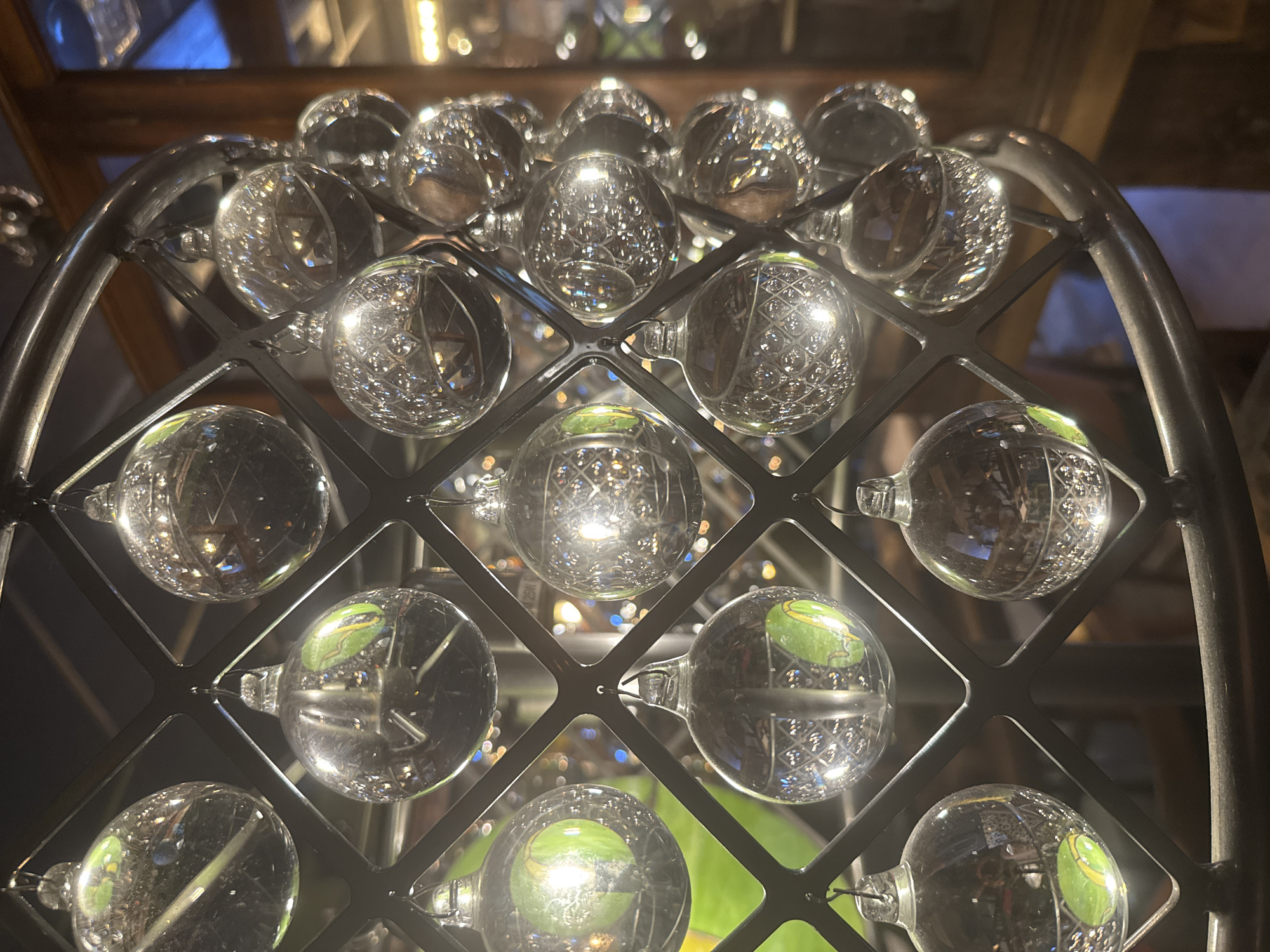 The image shows a close-up of a decorative light fixture, possibly a chandelier or a pendant light. It is comprised of a black metal frame with a lattice-like structure, from which numerous clear glass ornaments are suspended. These ornaments are spherical and reflect the surrounding environment, showing distorted images of lights, greenery, and possibly furniture. The background is blurred, but suggests an indoor setting with warm lighting and dark wooden elements. There are no people visible in the image. The specific location in Moscow, Russia, is not discernible from the visual information provided in the image.