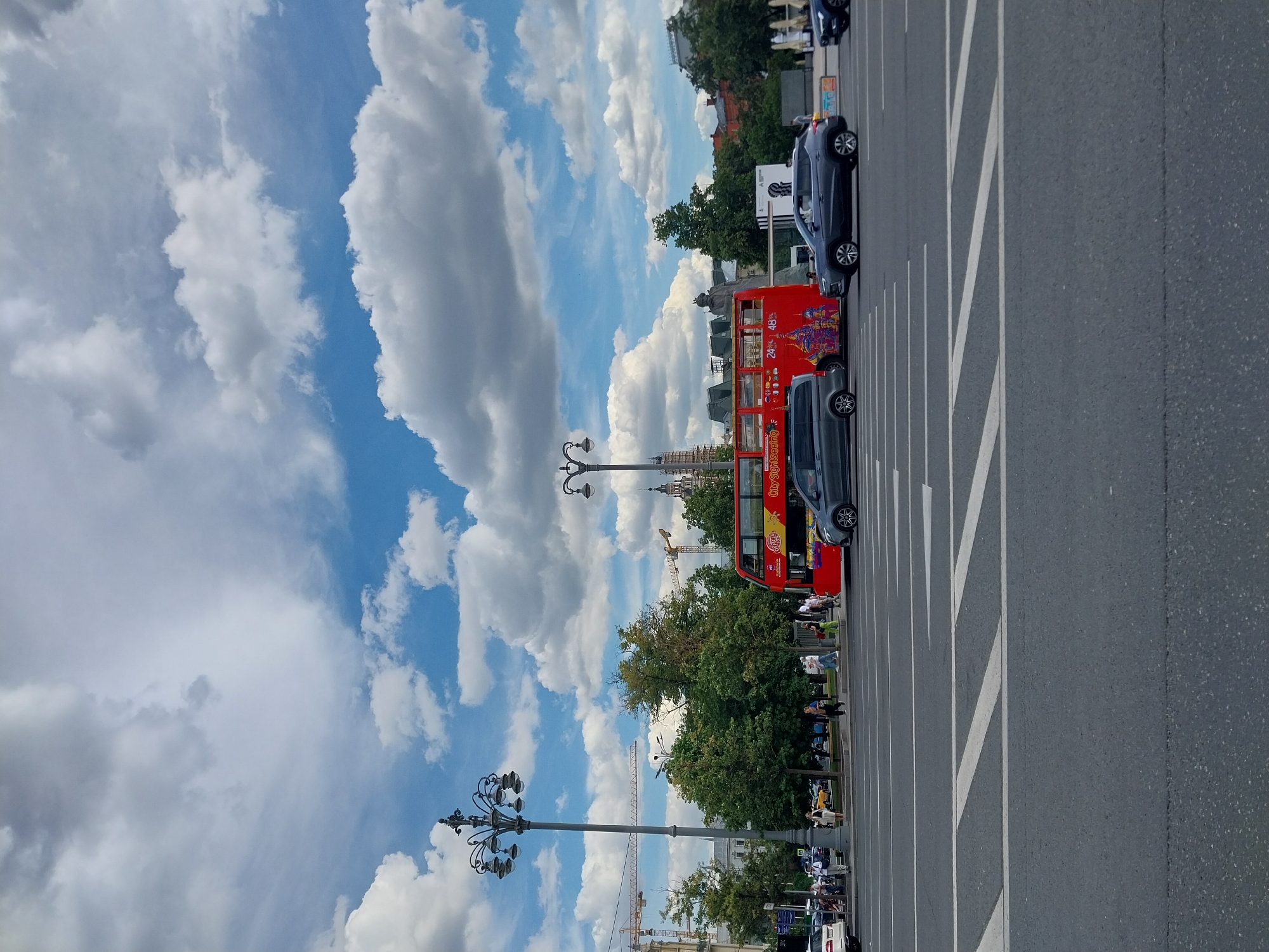 This image depicts a street scene in Moscow, Russia, likely during the daytime on a cloudy but bright day. The main subjects are a bright red double-decker tour bus and several cars parked or driving on a multi-lane asphalt road marked with white lines. In the background, there are trees, buildings, and cranes, suggesting an urban environment with ongoing construction or development.
Several people are visible on the sidewalk and near the bus, some appearing to be tourists. The sky is filled with dramatic, large white clouds against a blue backdrop, typical of a summer day. The time of day is likely late morning or early afternoon, given the brightness and the cloud formations.
The red tour bus has text on its side, including "24°" and "48°", likely indicating temperature or information relevant to the tour. There is also smaller text that appears to say "CitySightseeing" on the lower part of the bus, confirming its function as a tour vehicle. A dark-colored SUV is parked behind the bus, and another dark car is driving in the same lane.
Street lamps with multiple ornate fixtures are present on the sidewalk. The overall atmosphere seems to be one of normal city activity, with the presence of tourists and traffic. The image is oriented vertically, giving a sense of the height of the sky and the depth of the street.
