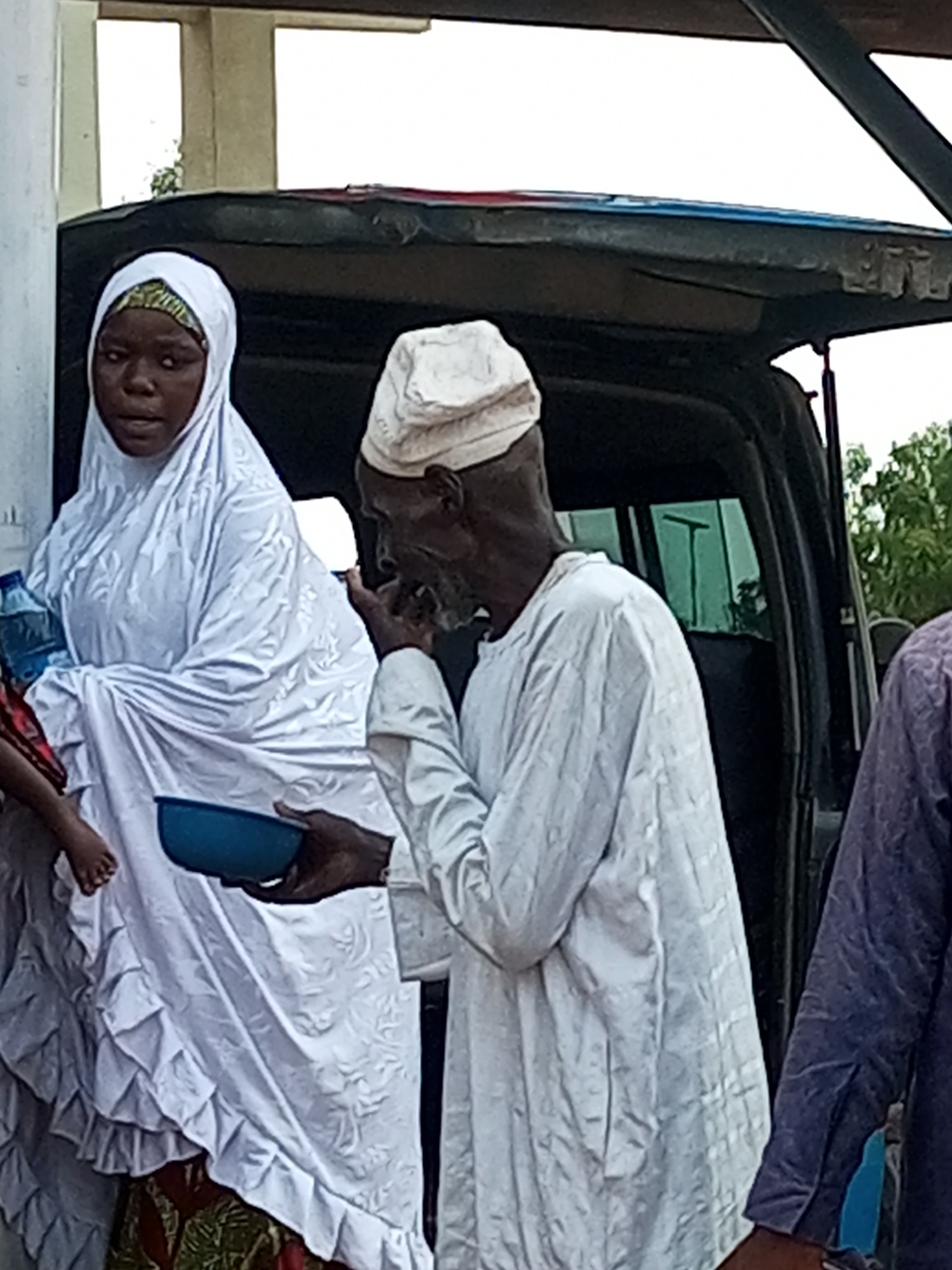In Gombe, Nigeria, an elderly man wearing a white cap and a long white robe is shown from the waist up, looking to his left. He is holding a blue bowl in his left hand and appears to be interacting with a woman in front of him. The woman, also seen from the waist up, is wearing a white hijab that covers her head and shoulders, and a patterned garment beneath. She is holding a blue plastic water bottle in her right hand. In the background, a dark-colored vehicle with an open back is visible. The scene appears to be outdoors, with some foliage in the far background. The overall impression is of a moment of exchange or offering between the two individuals.