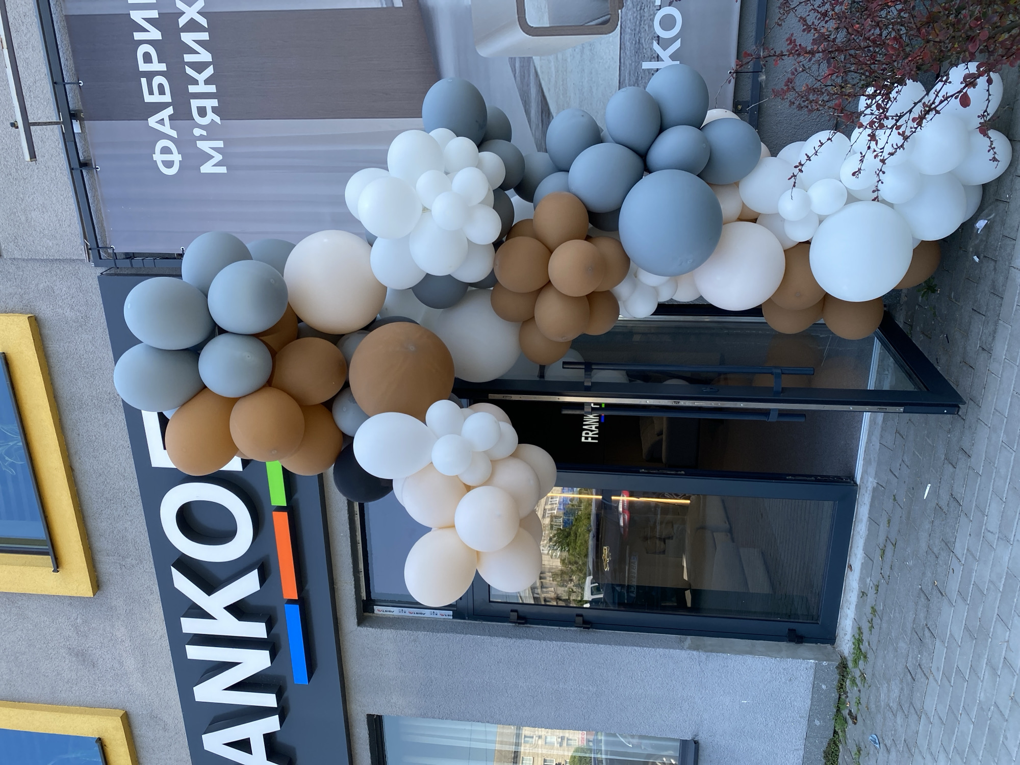 This is a medium shot, eye-level, exterior photograph of the entrance to a business named "ANKOF". The facade of the building is grey stucco. Above the entrance is a black sign with white, three-dimensional lettering spelling out "ANKOF". Below the lettering are three horizontal colored rectangles: blue, orange, and green. To the right of the "ANKOF" sign is a second sign, partially obscured by balloons, with white Cyrillic lettering that reads "ФАБРИКА М'ЯКИХ" which translates to "soft furniture factory". A large, colorful balloon arch, composed of balloons in shades of white, cream, brown, grey, and light blue, frames the entrance. The entrance itself is a glass door with a dark grey frame. The door is slightly ajar, revealing a glimpse of the interior, which appears to be a retail space with furniture. The ground in front of the entrance is paved with grey interlocking bricks. To the right of the entrance, a small bush with dark red leaves is visible. The image is taken on an overcast day.