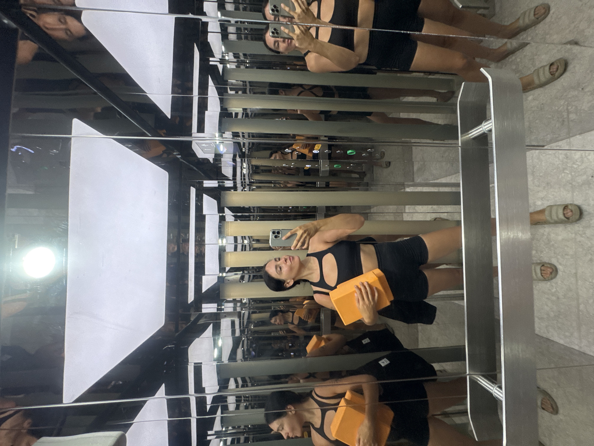 This is a vertical, eye-level, wide shot of a woman in an elevator with mirrored walls. The woman is facing away from the camera, taking a selfie in the mirror with her phone. She is wearing a black sports bra and black shorts. She is holding two orange yoga blocks. Her hair is dark brown and pulled back into a bun. She is wearing hoop earrings and a watch on her left wrist. In the reflection, we see the woman's face from the front, and she is looking down at her phone. Her expression is neutral.
The elevator has stainless steel walls and doors, and there are several horizontal lines of light on the walls. There are also elevator buttons visible in the reflection. The floor of the elevator appears to be made of polished concrete or stone tiles. In the upper part of the image, we see another reflection of the woman from the chest up, and she is wearing a black tank top. In the lower part of the image, there are reflections of the woman's legs and feet, and she is wearing a pair of light-colored slip-on sandals.
The overall impression is that of a woman taking a fitness-related selfie in a modern elevator. The lighting is bright and reflects off the mirrored surfaces, creating a dynamic and somewhat disorienting effect. The time of day is not discernible. The location is North Kuta, Indonesia. There are no visible emotions other than neutrality.