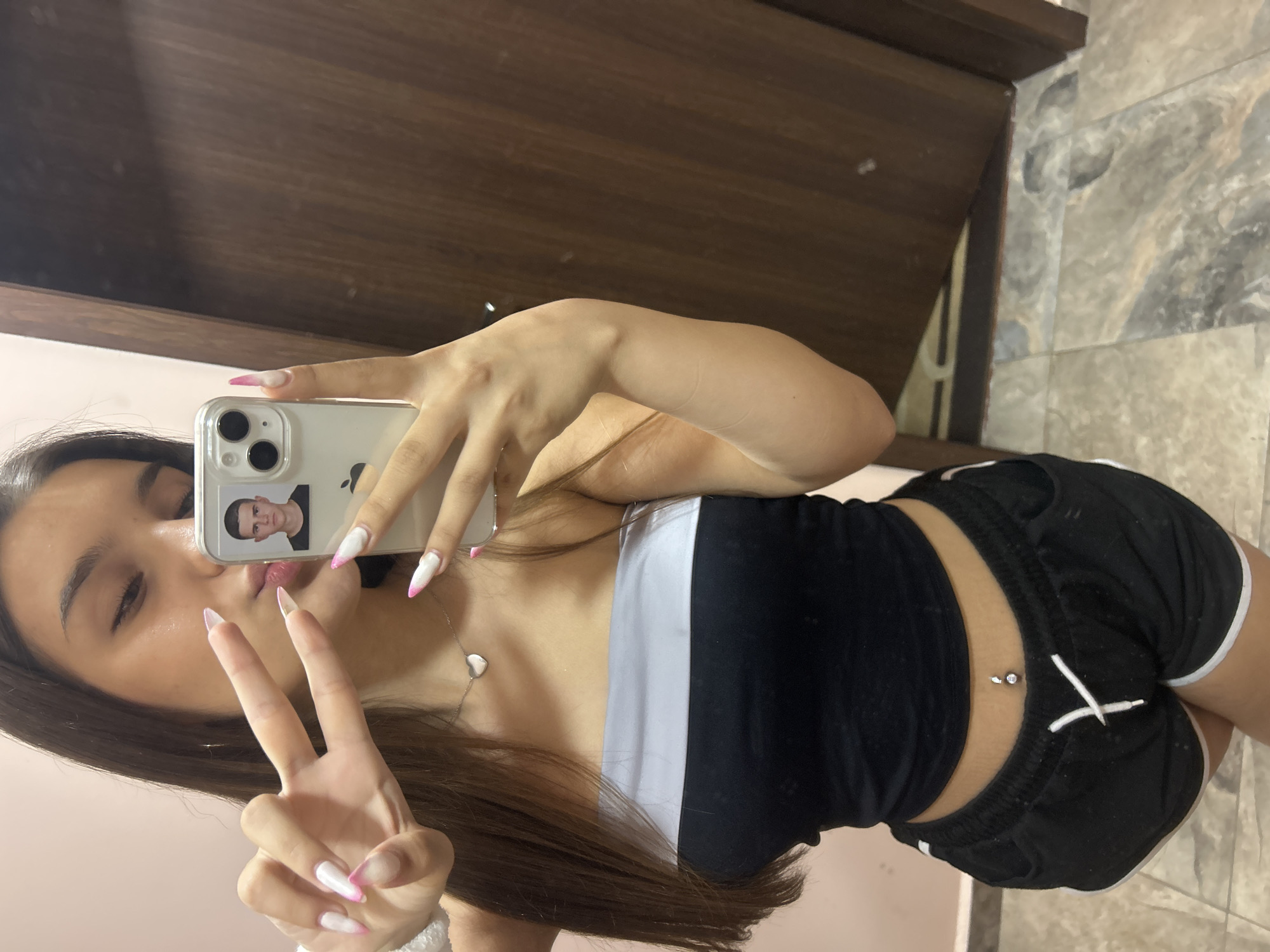 The image is a selfie taken by a young woman in a room. She is wearing a black and white strapless top and black shorts. She has long, dark hair and is wearing a heart-shaped necklace and a belly button piercing. Her fingernails are long and painted white with pink tips. She is holding up her hand in a peace sign, and a phone is visible in her other hand, showing a picture of a man. The background appears to be a wall and a wooden door. The flooring is tiled. The image is taken indoors, likely in a residential setting. The specific location in the city of Shumen, Bulgaria, cannot be determined from the visual information provided.
