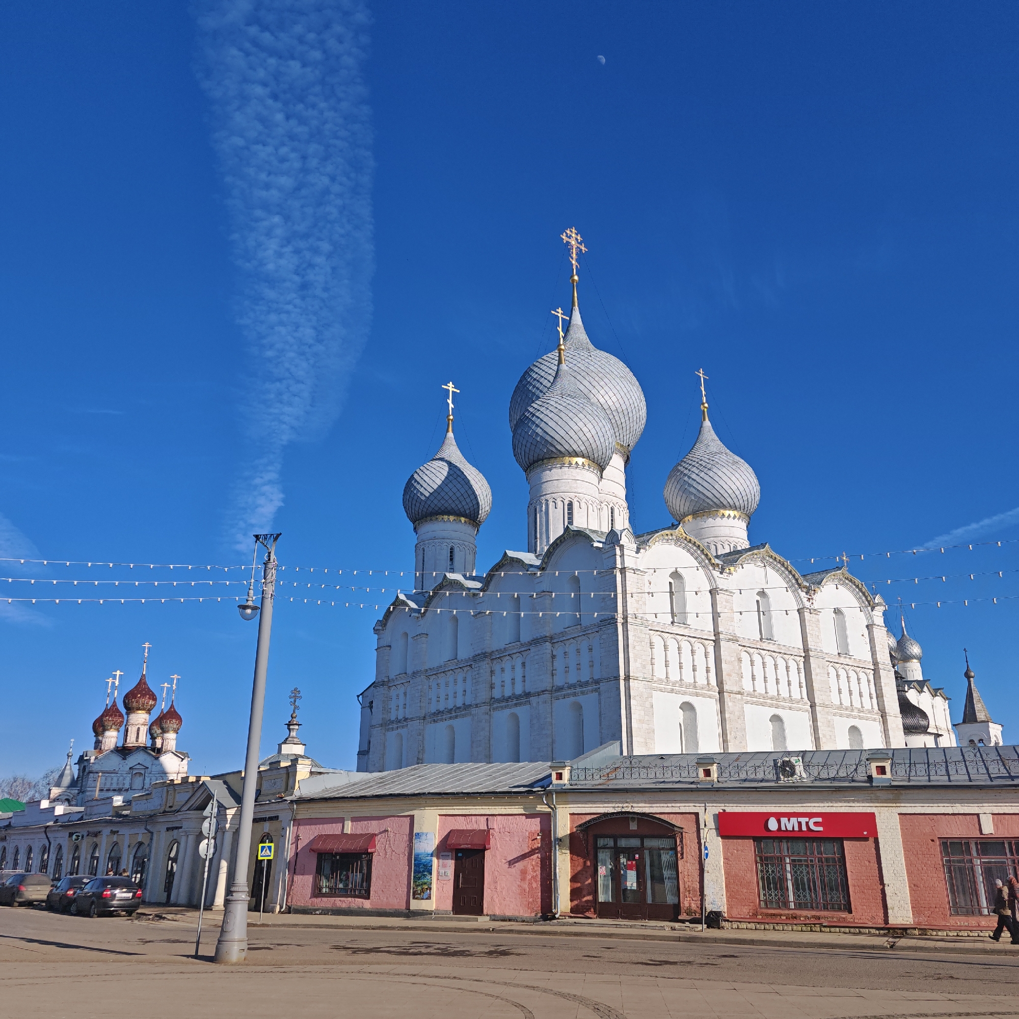 The image depicts a daytime scene in Rostov Veliky, Russia, featuring the architectural complex of the Rostov Kremlin.
**Sky and Atmosphere:** A clear, bright blue sky occupies the upper portion of the frame. A slender crescent moon is visible in the upper center-right. A faint, linear trail of cirrus clouds or a contrail extends vertically down the left side of the sky.
**Main Subject (Central):** The dominant structure is a large, white-plastered Orthodox cathedral, identifiable as the Dormition Cathedral (Uspensky Cathedral). It features five prominent, intricately textured, grey-silver onion domes, each topped with a golden Orthodox cross. The cathedral's walls exhibit multiple arched windows, decorative pilasters, and cornices.
**Left-Midground Structures:** To the left of the main cathedral, a smaller church building is partially visible. It features three reddish-brown, darker domes also topped with golden Orthodox crosses, suggesting it is part of the same historical complex. In front of this, a long, single-story building with a light-colored facade (cream/yellow) and multiple arched windows/doorways runs parallel to the street. Several dark-colored cars (at least two sedans) are parked along the street in front of this building.
**Foreground Structures (Right):** A row of single-story buildings runs across the lower portion of the image. The building closest to the viewer on the right features reddish-pink brick walls with a white base. One storefront has a prominent red "MTC" (MTS) logo above a barred window. Another storefront to its left has a dark brown door and a dark red awning.
**Street and Pedestrian Details:** The foreground consists of a paved street or square. A grey lamppost stands prominently in the mid-ground. Overhead, multiple strings of small, white decorative lights stretch horizontally across the frame. A blue-and-white pedestrian crossing sign (zebra crossing) is visible near the lamppost. On the far right sidewalk, a single individual, dressed in dark clothing, walks away from the camera.