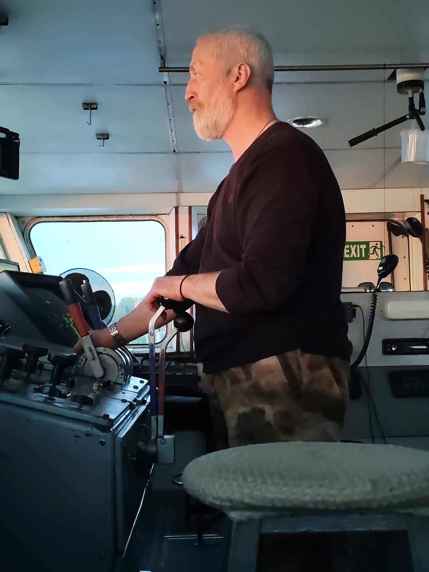 A medium shot depicts a man standing at the controls of what appears to be a ship's bridge. The man is facing to the left, his right hand holding onto a lever. He is wearing a dark long-sleeved shirt and camouflage-patterned pants. The man has gray hair and a beard. The bridge includes a variety of control panels and levers, as well as an "EXIT" sign. A small round stool is visible in the lower portion of the image. The location appears to be inside the control room of a ship, possibly navigating a waterway. No specific city or unknown location details are visible.