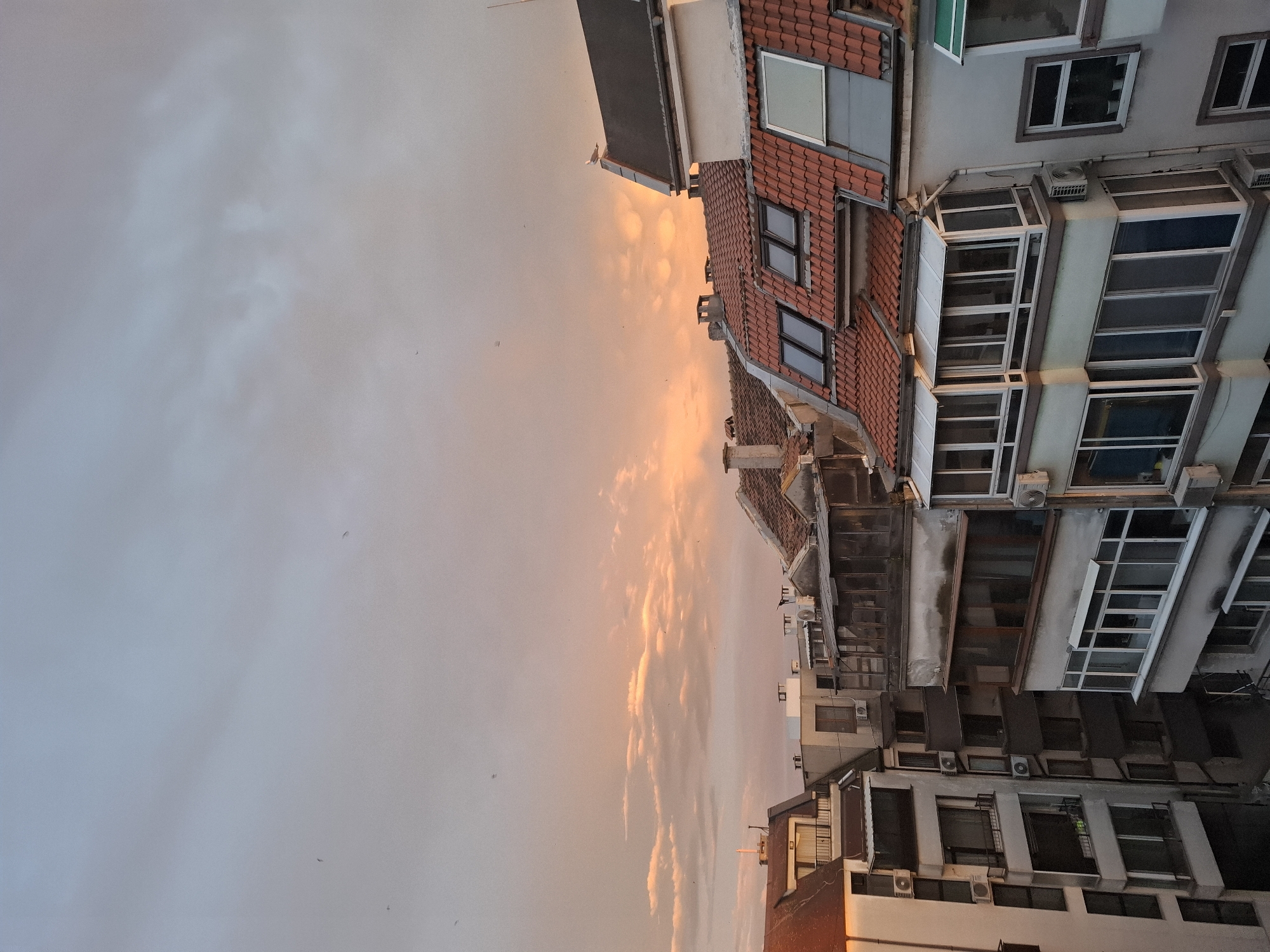 The image depicts a view of buildings and a sunset sky, possibly taken in Burgas, Bulgaria. The buildings are mostly residential, with a mix of architectural styles. Some have red tile roofs and windows with white frames. Air conditioning units are visible on some of the buildings. The sky is filled with clouds, illuminated by the colors of the setting sun.