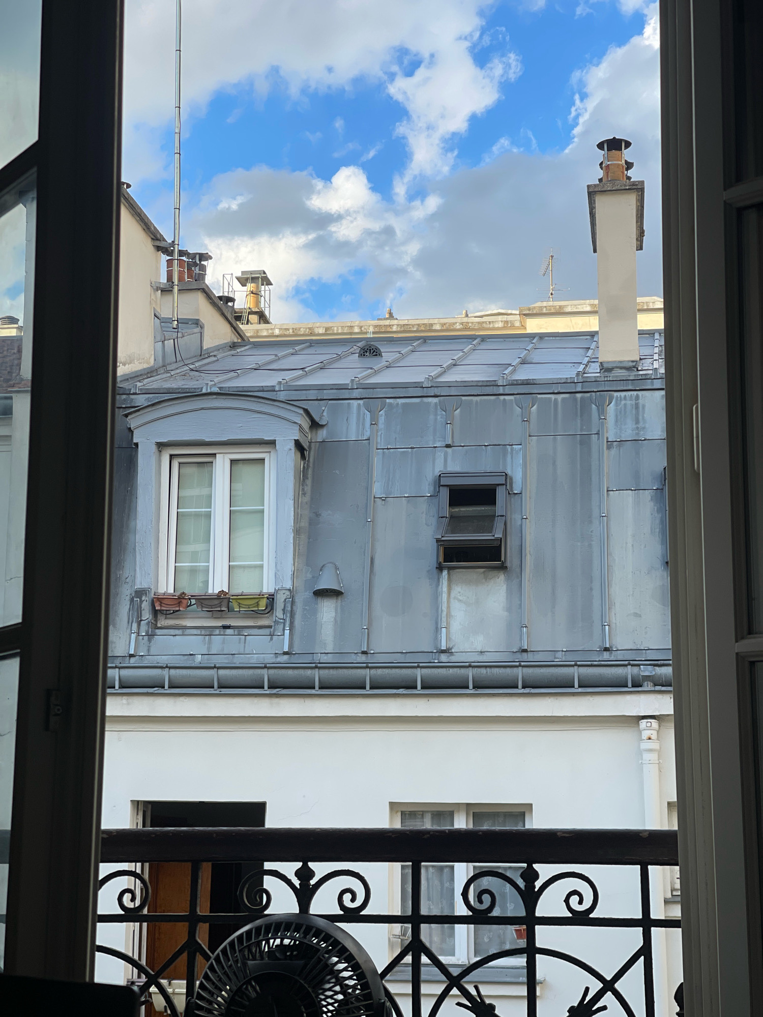 The image captures a view of Parisian rooftops from an open window, showcasing typical French architectural elements. The foreground is dominated by the interior of a room, with the edges of a window frame and a black metal railing visible. Beyond the railing, a white building with windows is seen, leading up to the iconic zinc-covered mansard roofs characteristic of Paris. Several chimneys, a common sight in the Parisian skyline, punctuate the rooftops. One prominent chimney is tall and rectangular, topped with a decorative cap. Another is shorter and made of brick. A small, open dormer window is visible on the zinc roof, suggesting an attic space. The sky above is a mix of blue with scattered white clouds, indicating daylight. A thin, tall metal pole, likely an antenna or lightning rod, extends vertically into the sky from one of the buildings. The overall impression is a classic urban scene in Paris.