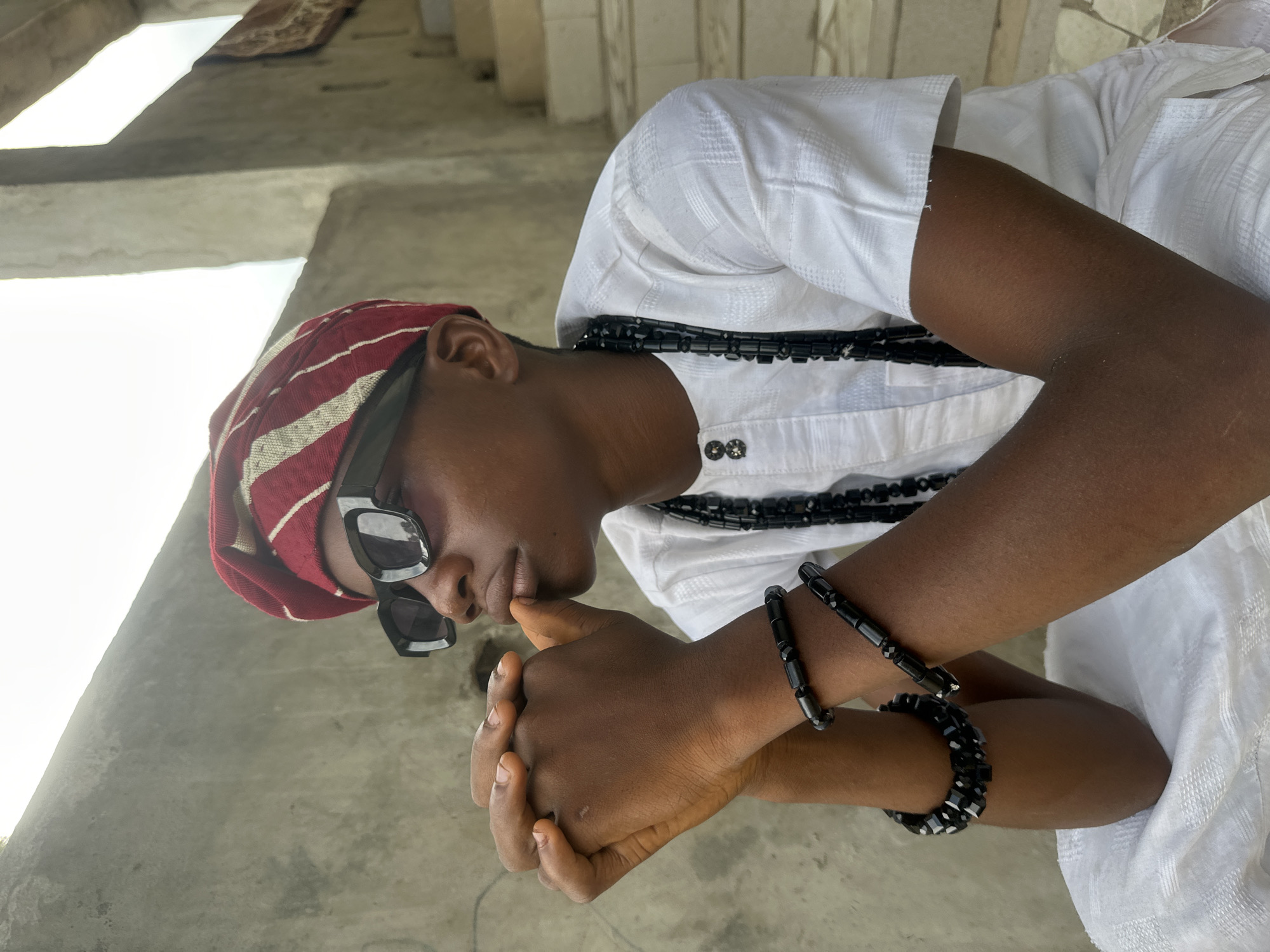 A young Black male is seated outdoors, looking to his right and resting his chin on his clasped hands. He wears a red and white striped headwrap, black sunglasses, a white embroidered tunic, and multiple black beaded necklaces and bracelets. The background consists of a textured concrete wall and a set of stone stairs with a patterned rug draped on the upper steps. The lighting suggests a bright, overcast day. This scene is located in Badagry, Nigeria, a city known for its historical significance as a former slave port, with many such architectural elements and outdoor settings common in the area.