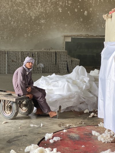 A man wearing traditional Afghan clothing, including a head wrap, sits on a small handcart in what appears to be a cotton processing or storage facility. The building has bare concrete walls that appear aged and possibly stained. Behind him, a large pile of white fabric or cotton sheets is visible. Further in the background, stacks of what look like cardboard or fiberboard dividers are arranged. The floor is concrete with loose cotton fibers scattered around. In the foreground, a red metallic object is seen, potentially part of machinery used in cotton processing. The scene suggests a textile-related industry setting. There is no other evidence suggesting the location is in Kandahar.