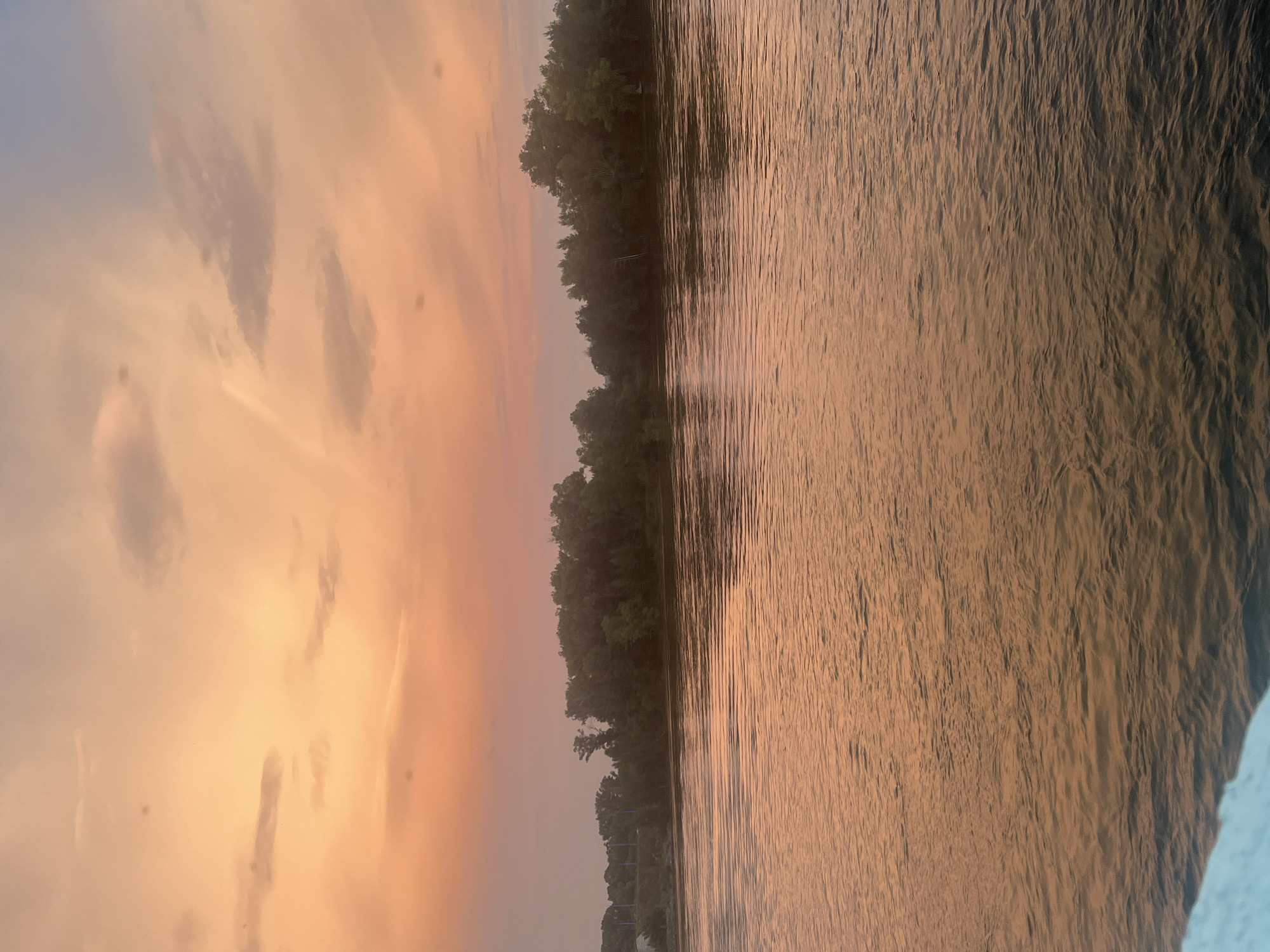 An image shows a river at sunset, with trees lining the far bank. The sky is a gradient of orange and pink, with scattered clouds. The water is reflecting the colors of the sky, creating a shimmering effect. The scene appears to be peaceful and serene. The image is taken from a low angle, suggesting the photographer is on a boat or the riverbank. The trees are dense and green, with some taller trees scattered among them. In the distance, there are some structures that could be part of a city or industrial area. The overall impression is of a natural landscape at the end of the day.