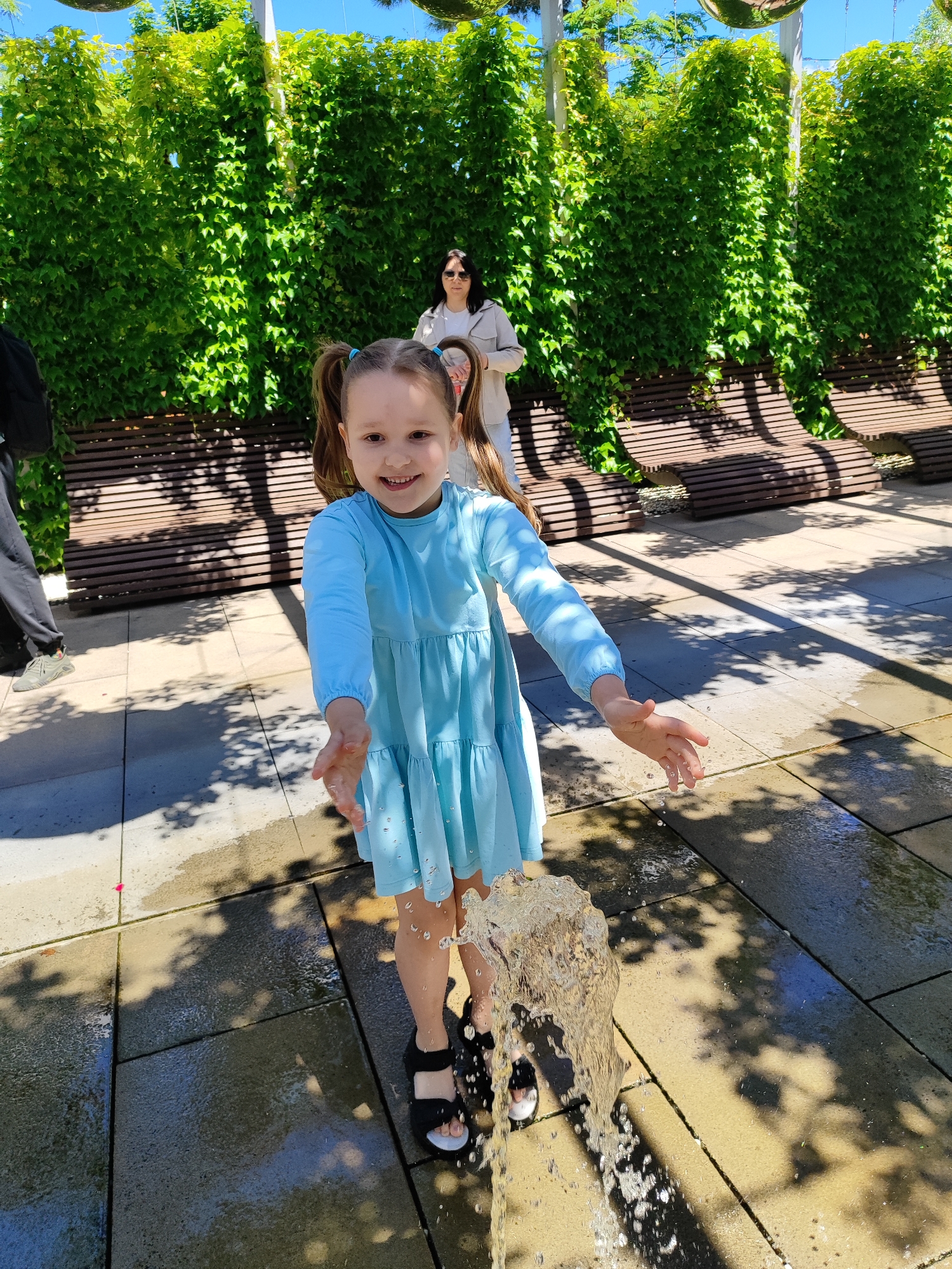 A young girl with pigtails, wearing a light blue tiered dress, is captured in a moment of joyful play with a gushing water fountain in an outdoor setting. She extends her hands into the upward jet of water, a wide smile lighting up her face as droplets splash around her. Her dark sandals are visible near the base of the fountain on the paved ground.
In the background, an adult woman with dark hair, wearing a light-colored jacket and sunglasses, stands observantly. A partially visible figure, likely another person, is on the far left of the frame. The scene is set in what appears to be a well-maintained park or urban garden, characterized by a tall, lush green wall of ivy or climbing plants that forms a backdrop. Several modern, slatted wooden benches are arranged behind the greenery. The ground is paved with light-colored tiles, marked by distinct, dappled shadows from overhead foliage or structures.
The image depicts a bright, sunny daytime, with clear blue sky visible at the top. The strong sunlight casts crisp shadows, indicating a clear weather day. The atmosphere is one of lightheartedness and amusement, centered around the girl's playful interaction with the water. No visible text is present in the image. This scene takes place in Krasnodar, Russia.