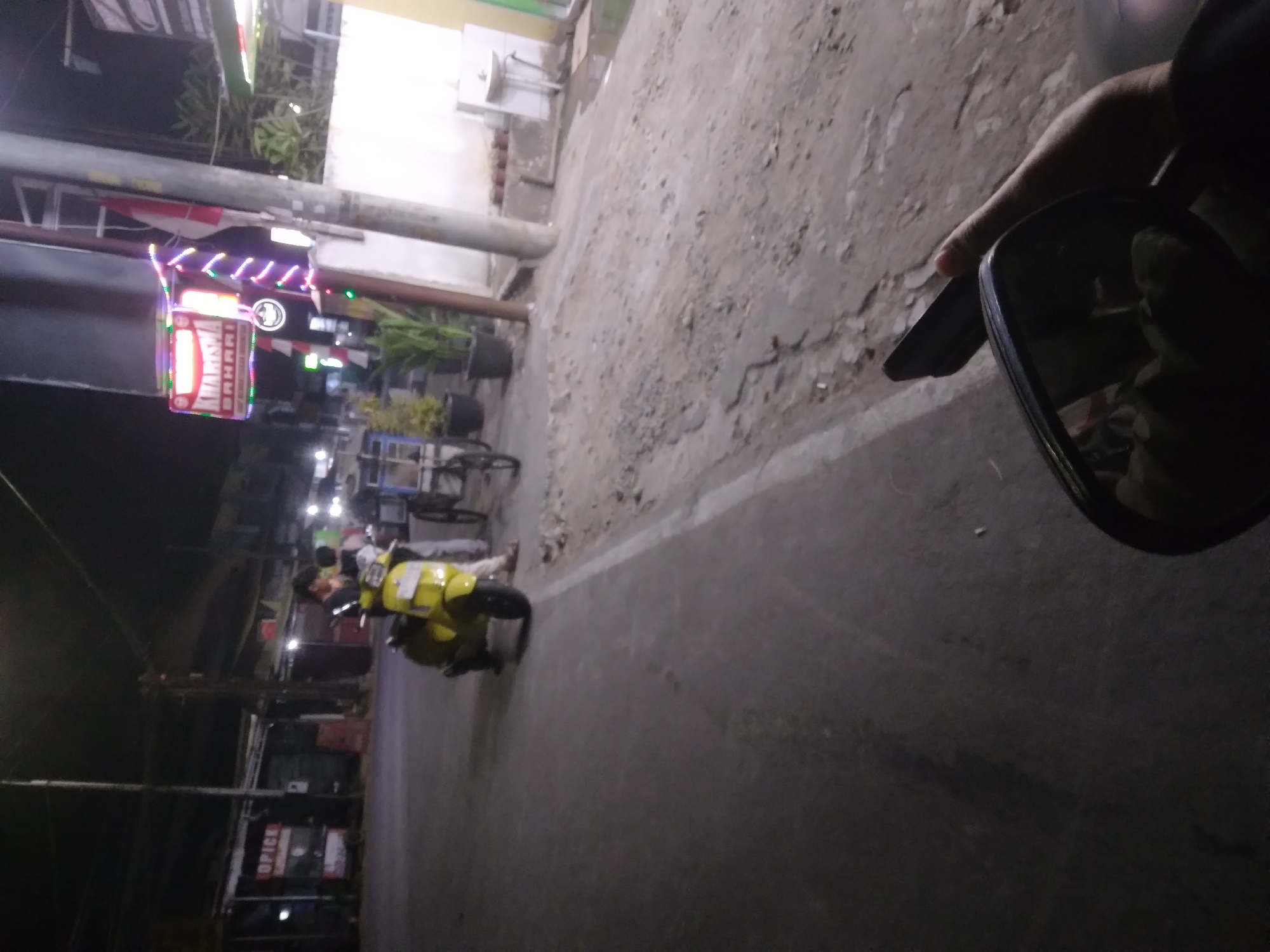 In Desa Pamijen, Kecamatan Sokaraja, Indonesia, a nighttime scene unfolds on a street. A person is seen standing next to a bright yellow scooter, which is parked on the left side of the asphalt road. Across the street, a sign with colorful neon lights that reads "KARISMA BAHARI" is illuminated. Further down the road, there's a sink and faucet on a pedestal, suggesting a public restroom or washing area. The environment is dimly lit, characteristic of a street at night, with some buildings visible in the background. The perspective suggests the image was taken from a moving vehicle, possibly on a motorcycle, as a side-view mirror is visible in the foreground.