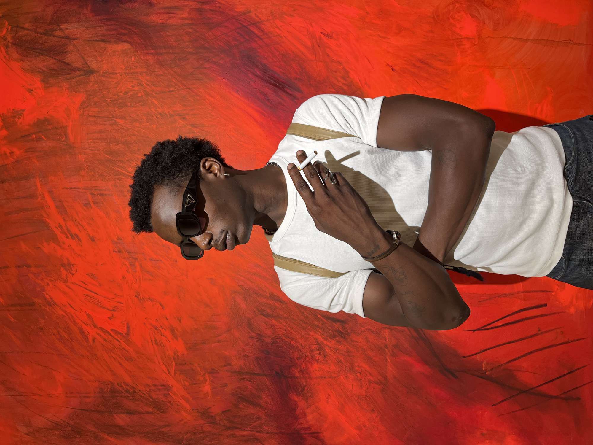 A young Black man with short, curly dark hair and sunglasses stands against a vibrant, textured red background. He is wearing a white t-shirt, dark denim jeans, and a light-colored strap over his shoulder. His arms are crossed, and he holds a lit cigarette between his fingers, bringing it to his lips. A silver ring is visible on his right hand. The red background appears to be a painting or mural with visible brushstrokes and varying shades of red and hints of black. The lighting suggests an indoor setting. The man is positioned slightly off-center to the right of the frame, looking to his left. There is no discernible specific location within Barcelona, Spain, that can be identified from the provided image.