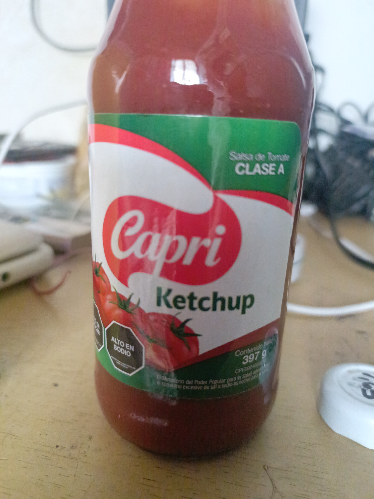 The image shows a bottle of Capri brand ketchup. The label is white with a red and green banner at the top. The word "Capri" is written in red script, and below it is the word "Ketchup" in black. There are images of tomatoes on the label. A black octagon on the left side of the label says "ALTO EN SODIO", indicating that the ketchup is high in sodium. Underneath the tomatoes on the right side of the label the label says "Contenido Neto 397 g". The bottle is sitting on a wooden surface. A white bottle lid, along with other various objects can be seen in the background. There are no people present in the image. The text "Salsa de Tomate CLASE A" is present on the label, which is in Spanish.