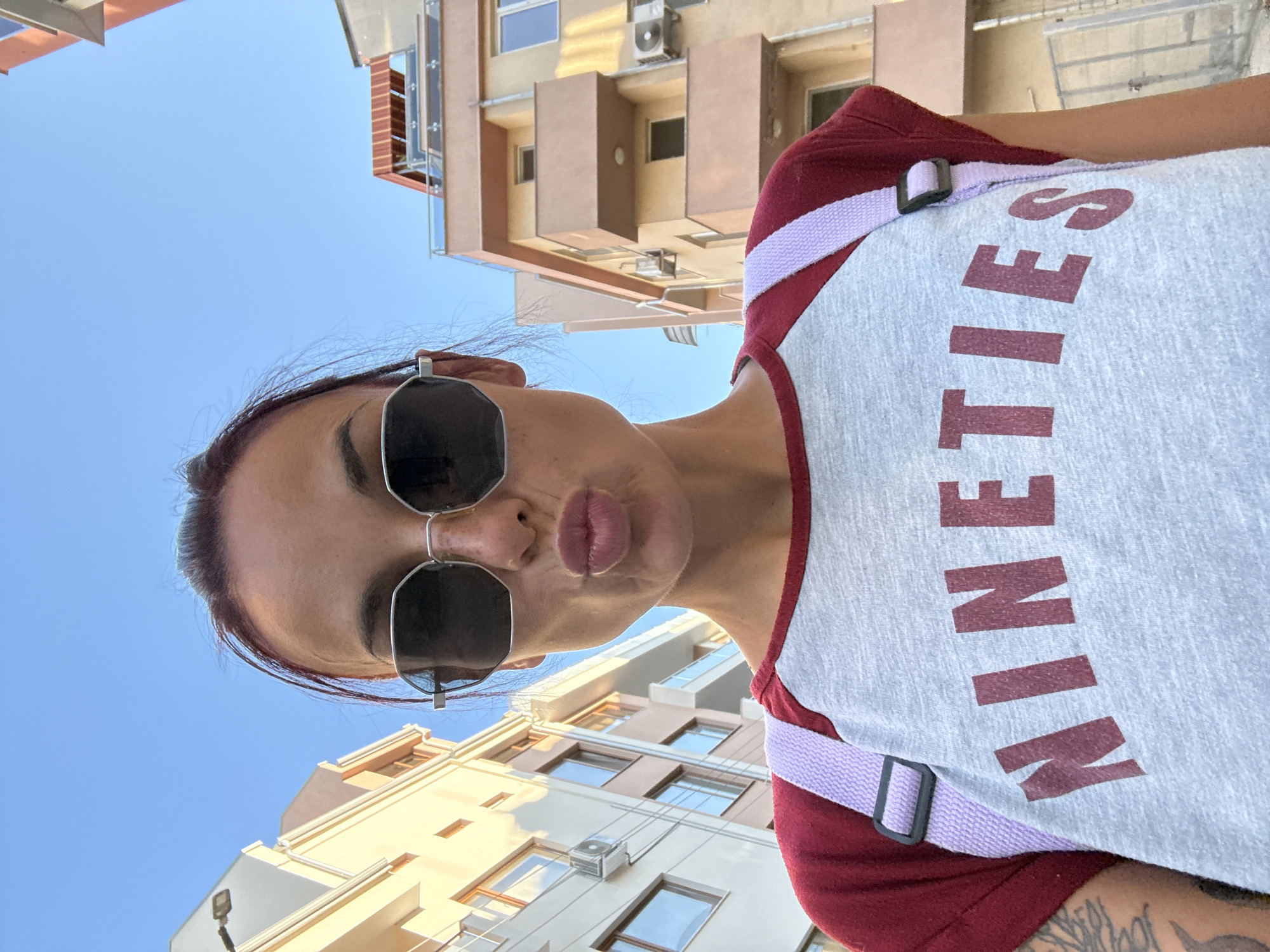 A young woman with dark hair pulled back and large, dark sunglasses is posing for a selfie on a city street. She is wearing a gray t-shirt with a burgundy neckline and sleeves, featuring the word "NINETIES" printed in burgundy letters. A light purple strap from a backpack is visible crossing her chest. She is puckering her lips for the photo. The background shows multi-story buildings with balconies and windows, indicative of an urban environment. The image appears to be taken during the daytime with a clear blue sky. The location is identified as the city of Pleven, Bulgaria.