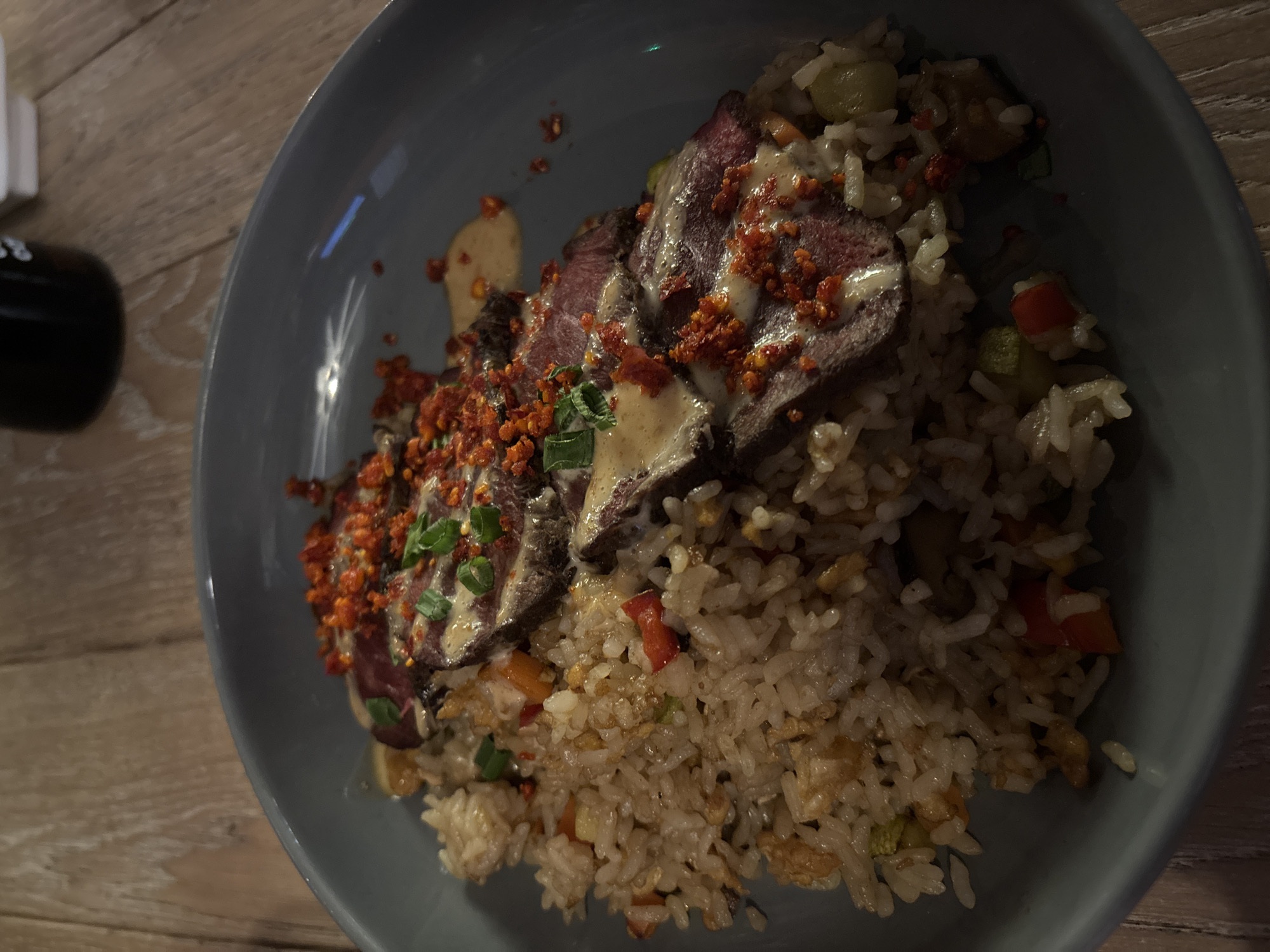 A close-up, overhead shot captures a visually rich meal served in a deep, light blue or grey plate, resting on a wooden table. The primary subject is a dish composed of sliced meat and fried rice.
The meat, appearing to be rare or medium-rare beef, is thinly sliced and arranged in a fan-like pattern on one side of the plate. It is generously drizzled with a creamy, light-colored sauce and heavily garnished with vibrant reddish-orange flakes, possibly chili crisps or a spiced crispy topping. Finely chopped green onions are scattered across the meat, adding a fresh green contrast.
Beside and beneath the meat lies a substantial portion of fried rice. The rice is mixed with various colorful ingredients, including diced red bell pepper, small green vegetable pieces (possibly peas or zucchini), and specks of what appears to be scrambled egg. The overall texture of the rice appears slightly glistening, indicating it's well-cooked and seasoned.
The lighting in the image is dim, suggesting an indoor setting, likely evening. This low light creates subtle reflections on the plate and enhances the rich colors of the food, particularly the red flakes and the deep red of the meat. On the far left, partially visible on the wooden surface, are indistinct outlines of other dining accessories: a dark, possibly cylindrical object and a white, rectangular object. No people or legible text are visible in the frame. The scene portrays a ready-to-eat meal, emphasizing its appetizing presentation.