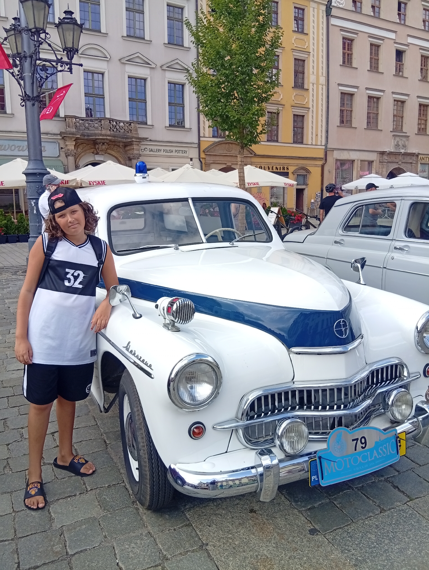 A young person stands next to a white and blue vintage car in a city square. The person, a boy with curly brown hair, is wearing a white sleeveless shirt with black trim and the number "32" in black, black shorts, and black slide sandals. He is wearing a black baseball cap backwards. The car is a white sedan with a blue stripe down the hood and a blue light on the roof. A license plate on the front of the car reads "79 MOTOCLASSIC". The car is parked on a cobblestone street in front of several buildings. The buildings are old and have multiple stories with arched windows. There are white umbrellas and some red flags visible in the background. The scene appears to be outdoors on a sunny day. The location is identified as Wrocław, Poland.