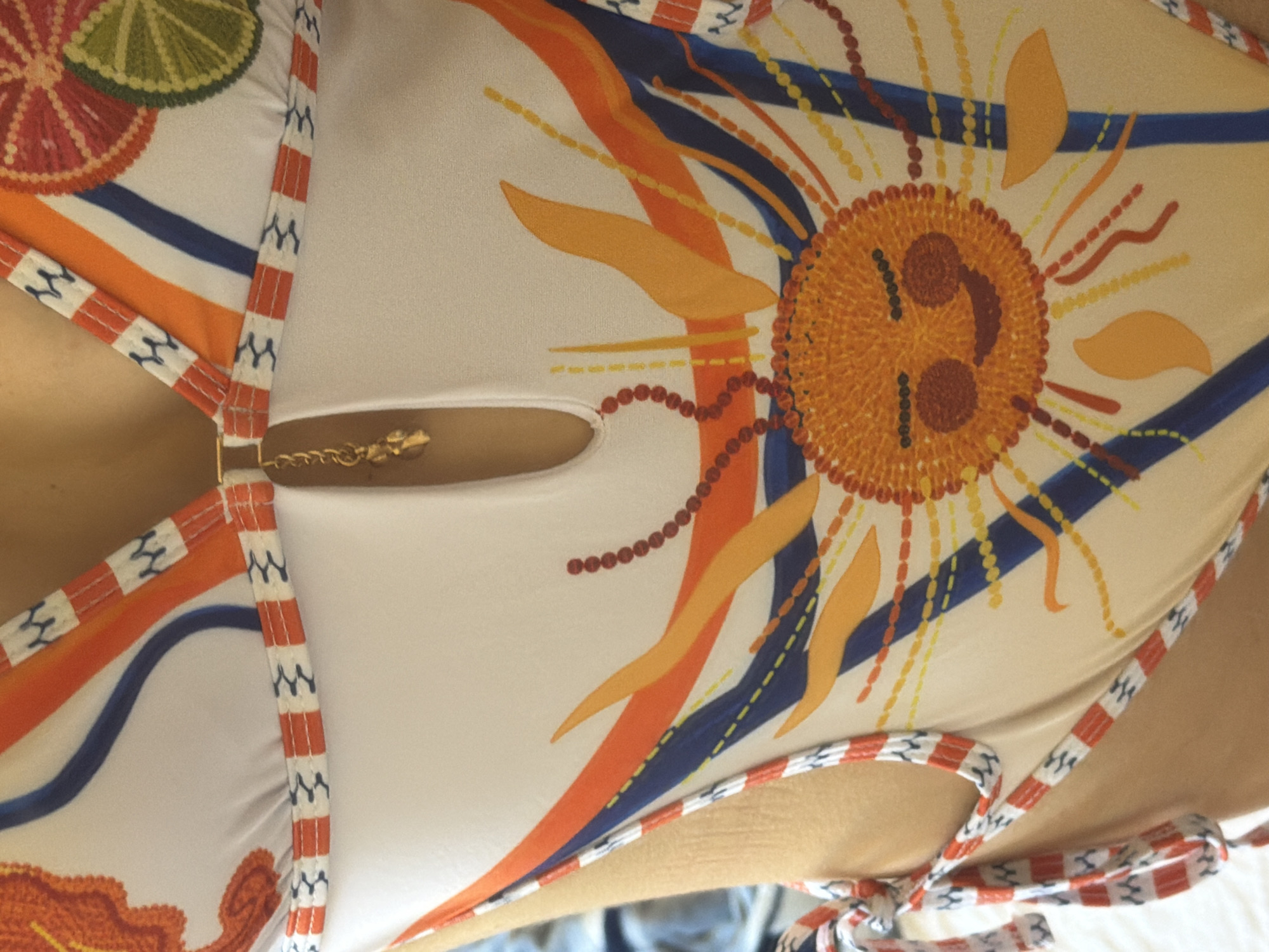 A close-up, low-angle, eye-level, medium shot captures a portion of a woman's torso wearing a one-piece swimsuit. The swimsuit is white with an all-over print featuring abstract orange, blue, and yellow designs, as well as circular fruit slices, possibly limes and oranges. A gold charm hangs from the neckline. The woman's skin is visible on her sides and back, with straps of the swimsuit tied around her shoulders. The background is out of focus and appears to be a light-colored surface, possibly bedding or a wall. The image is taken indoors, likely in a bedroom or dressing area, within the city of Tersefanou, Cyprus.