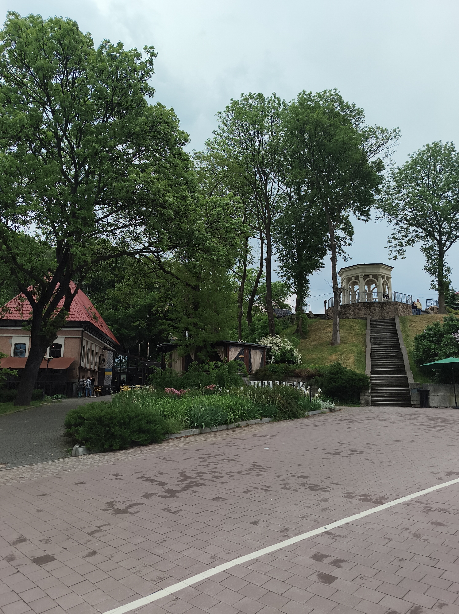 The image captures a view in Ternopil, Ukraine, under an overcast sky. A brick-paved area in the foreground has a white line marking. To the left is a building with a red-tiled roof and a group of people standing near the entrance. Adjacent to the building is a garden with flowers and greenery. In the background, a staircase leads up a grassy hill to a white gazebo. Trees surround the gazebo, partially obscuring it. A few people are present near the gazebo. A green umbrella and a trash bin stand to the right of the staircase. The environment suggests a park or recreational area.